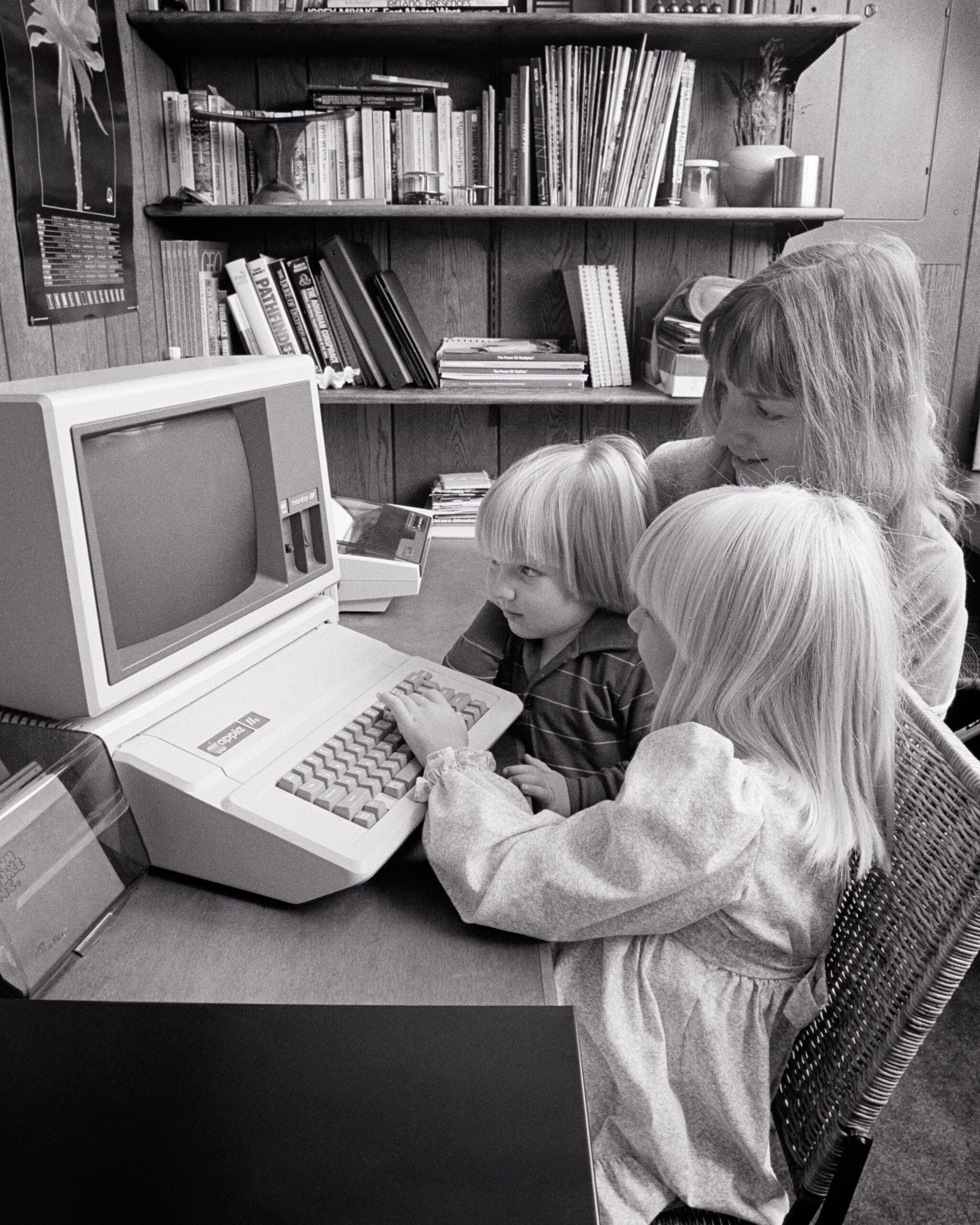 A woman and two children sit at a desk using an old Apple computer. Bookshelves and wood-paneled walls are in the background. The children are focused on the keyboard.