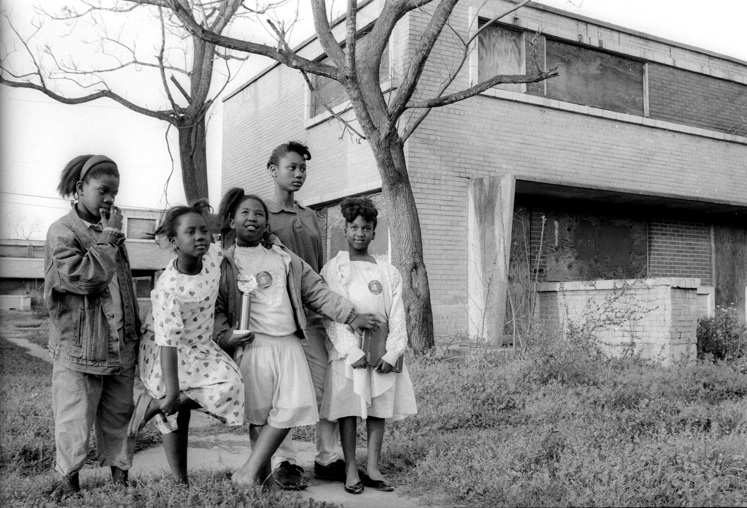 Five young girls stand together outside a brick building with boarded windows and bare trees. The girls are dressed in casual and semi-formal clothes, and the atmosphere feels candid and natural.