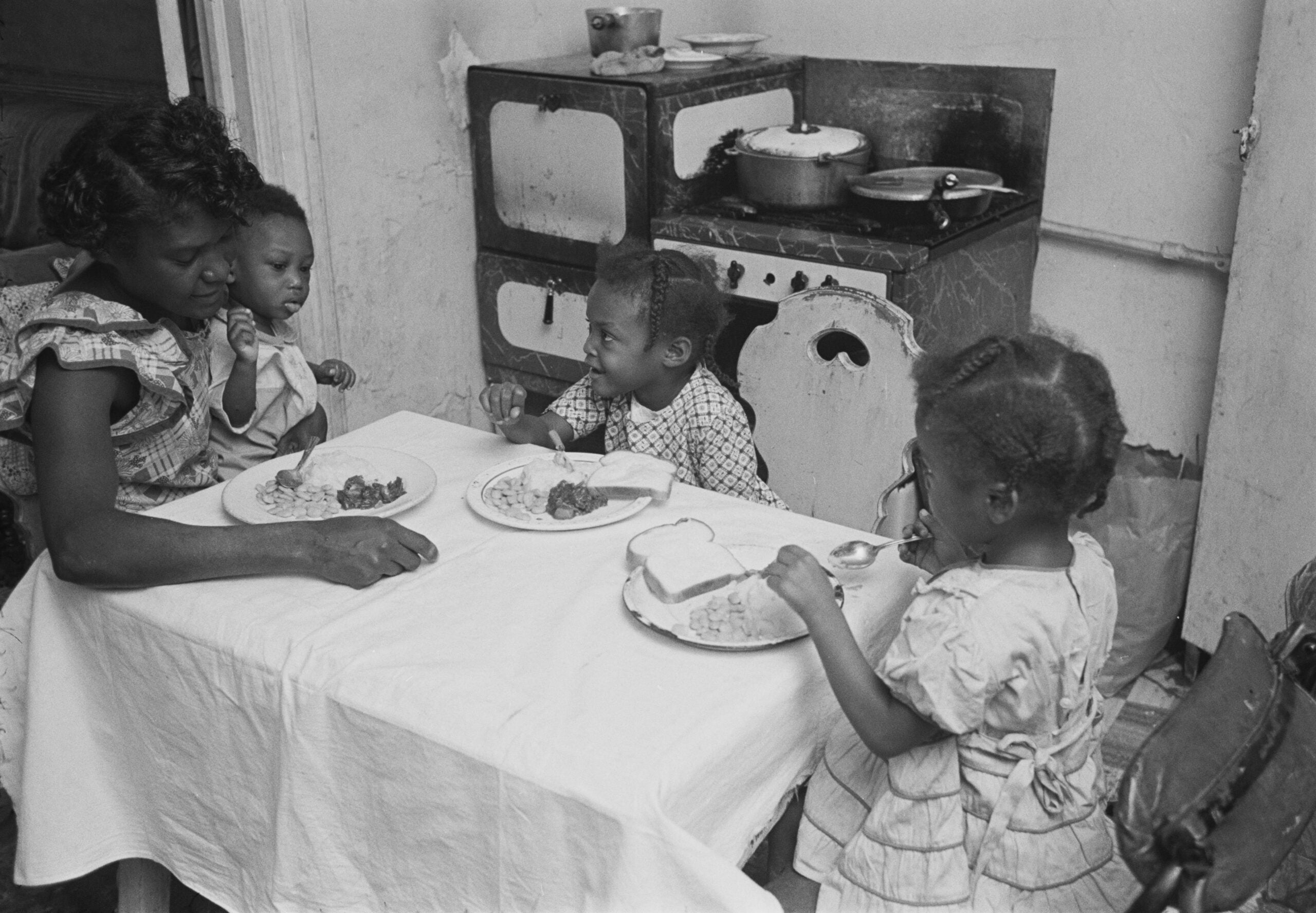 A woman and three young children sit at a small table, eating a meal together in a modest kitchen with pots on the stove in the background.