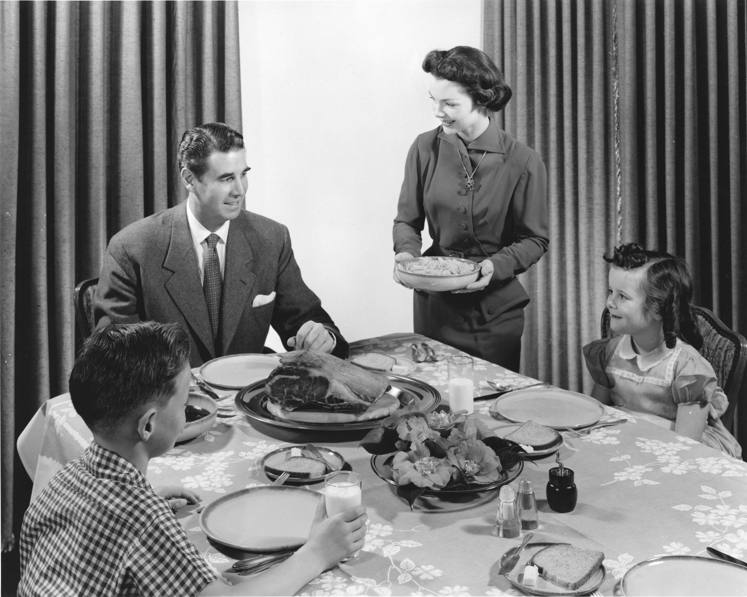 A woman serves pie to a man and two children seated at a dining table set with food and drinks. Everyone appears to be smiling and enjoying a family meal together.