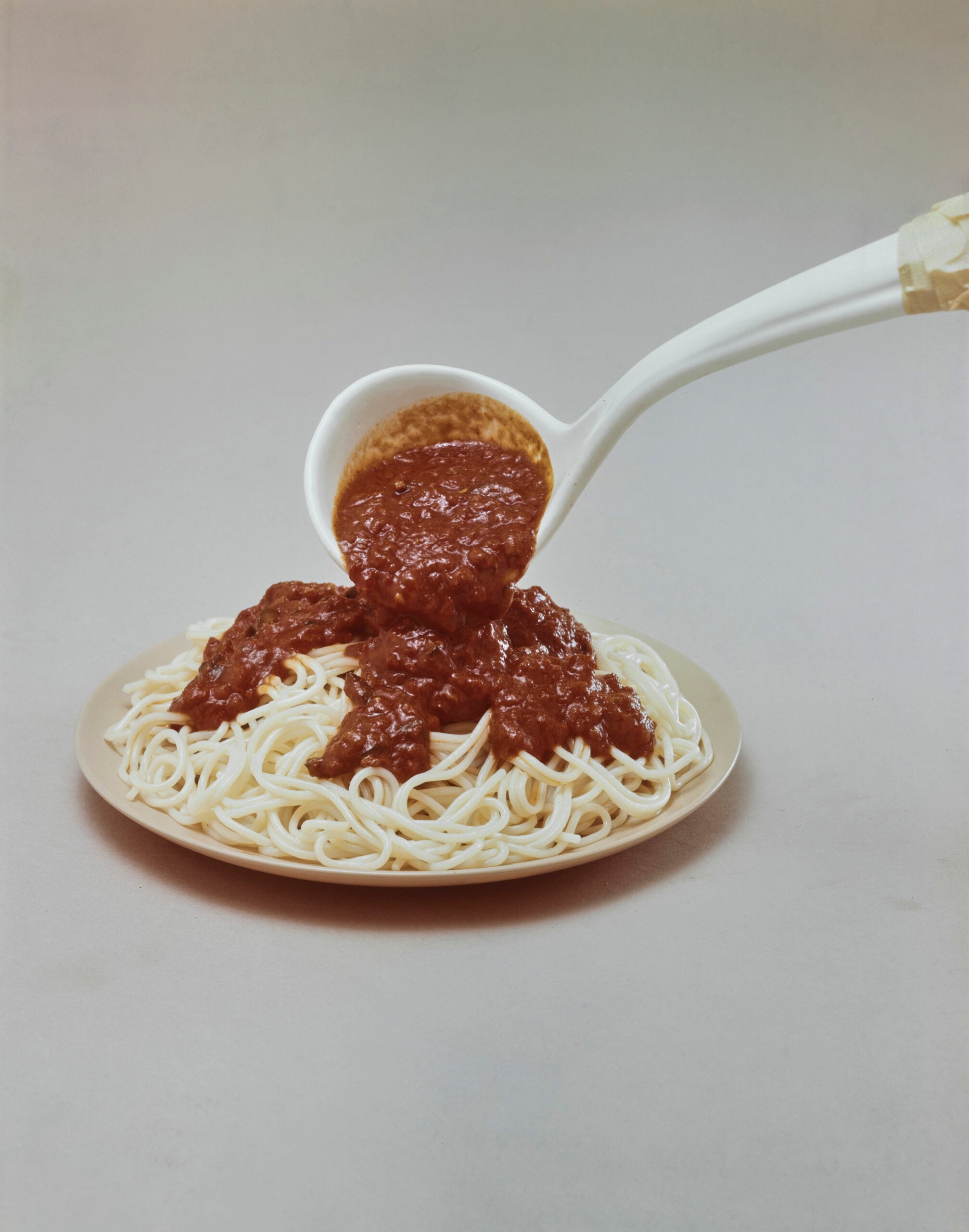 A white ladle pours chunky red tomato sauce over a plate of plain spaghetti on a neutral background.