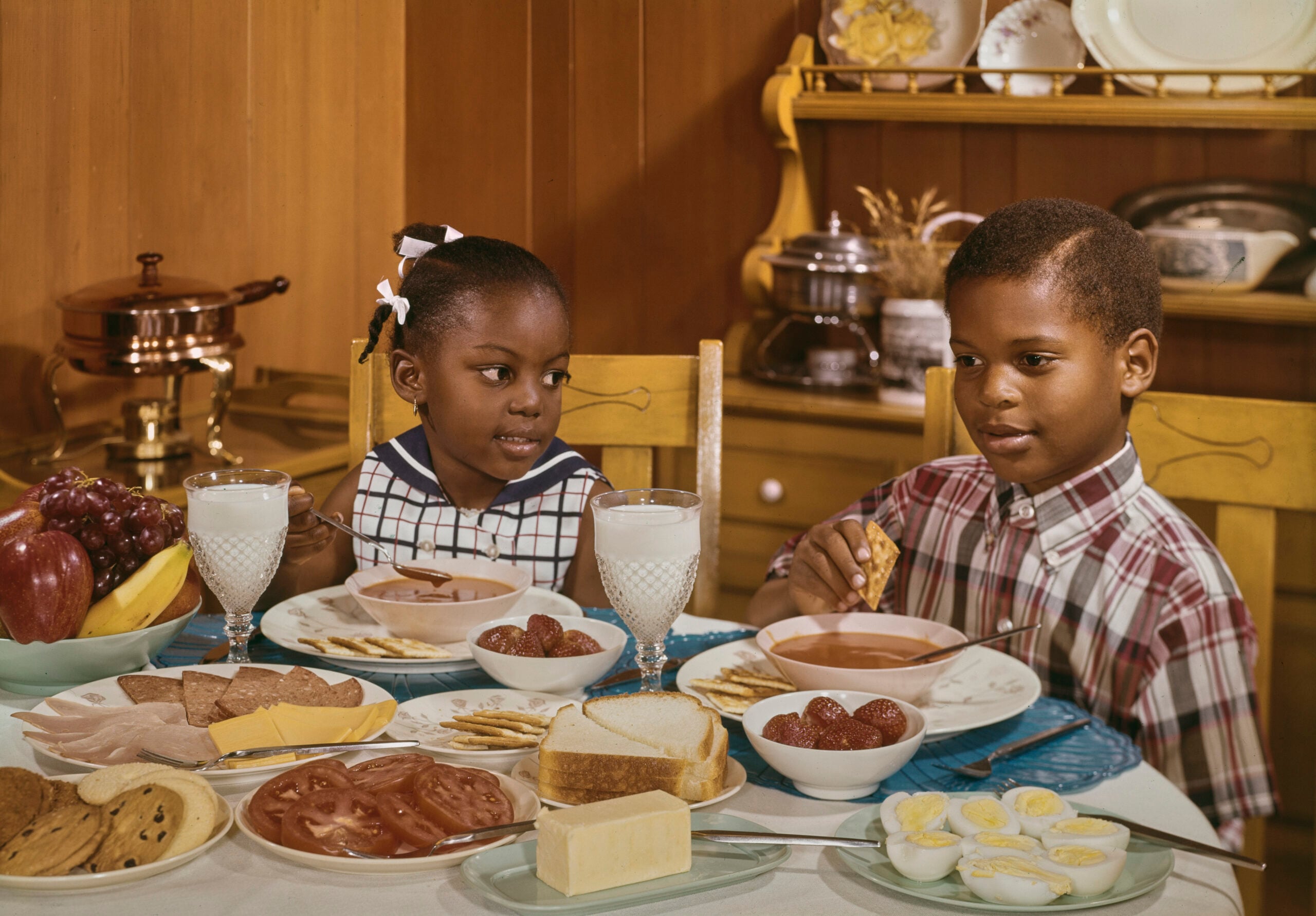 Two kids sit at a table with plates of soup, sliced bread, eggs, cookies, fruit, and glasses of milk. They are looking at each other in a cozy dining room with wooden walls and a hutch in the background.