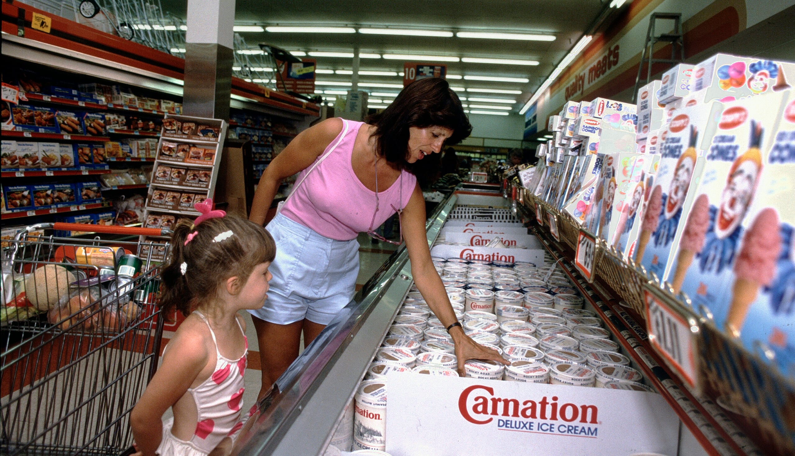 A woman in a pink top and a young girl shop for Carnation ice cream at a supermarket, standing by the freezer aisle next to a shopping cart and colorful boxes.
