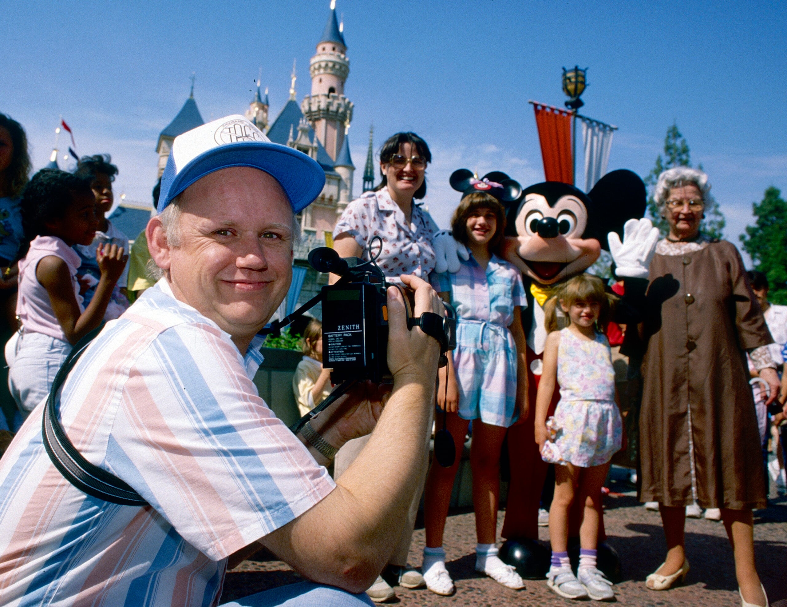 A man with a video camera smiles at the front, while a family poses with a person in a Mickey Mouse costume in front of a castle at a theme park on a sunny day.