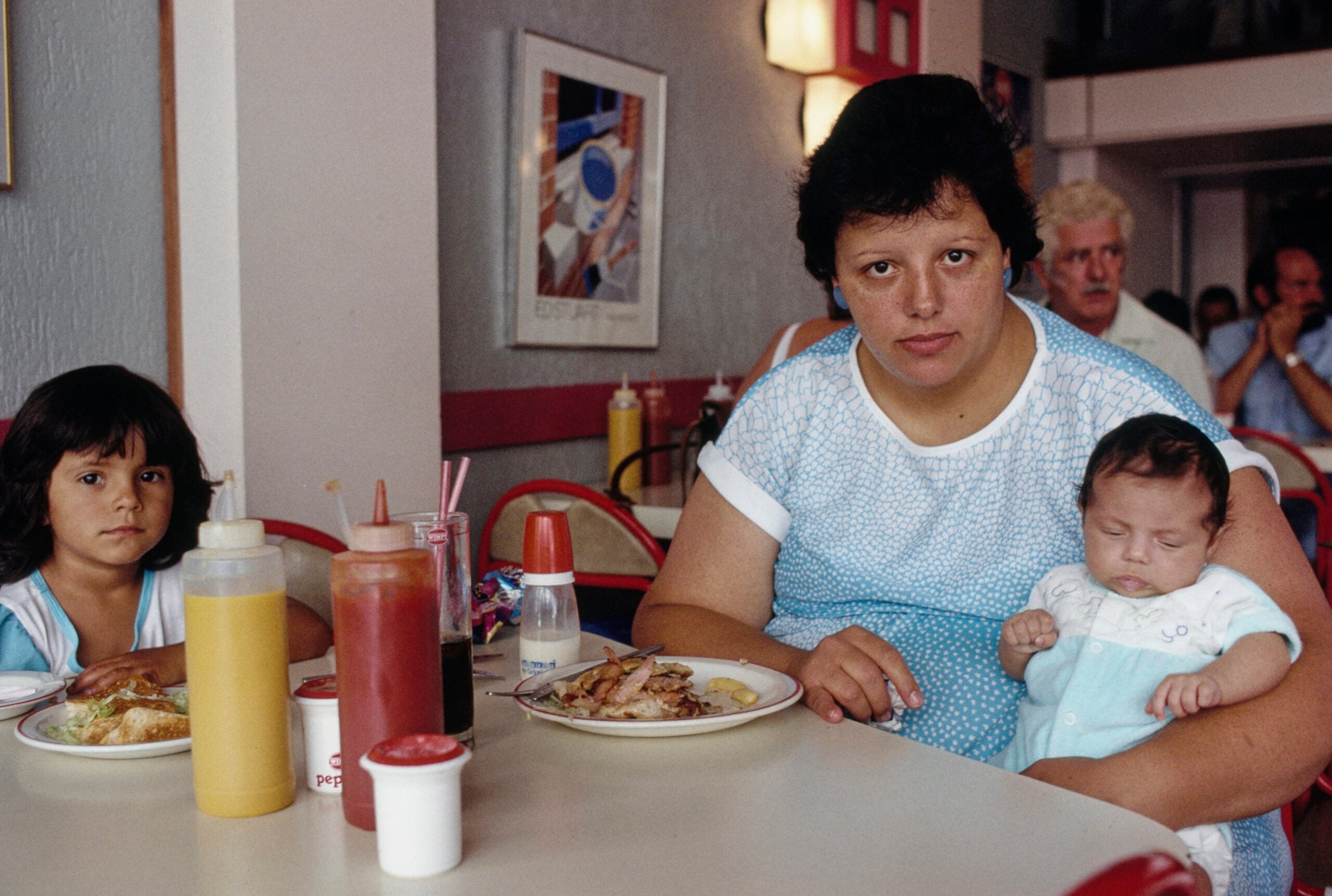 A woman holding a baby sits at a diner table with a young girl. Food, condiments, and a bottle are on the table. The woman looks at the camera; people and colorful decor are in the background.