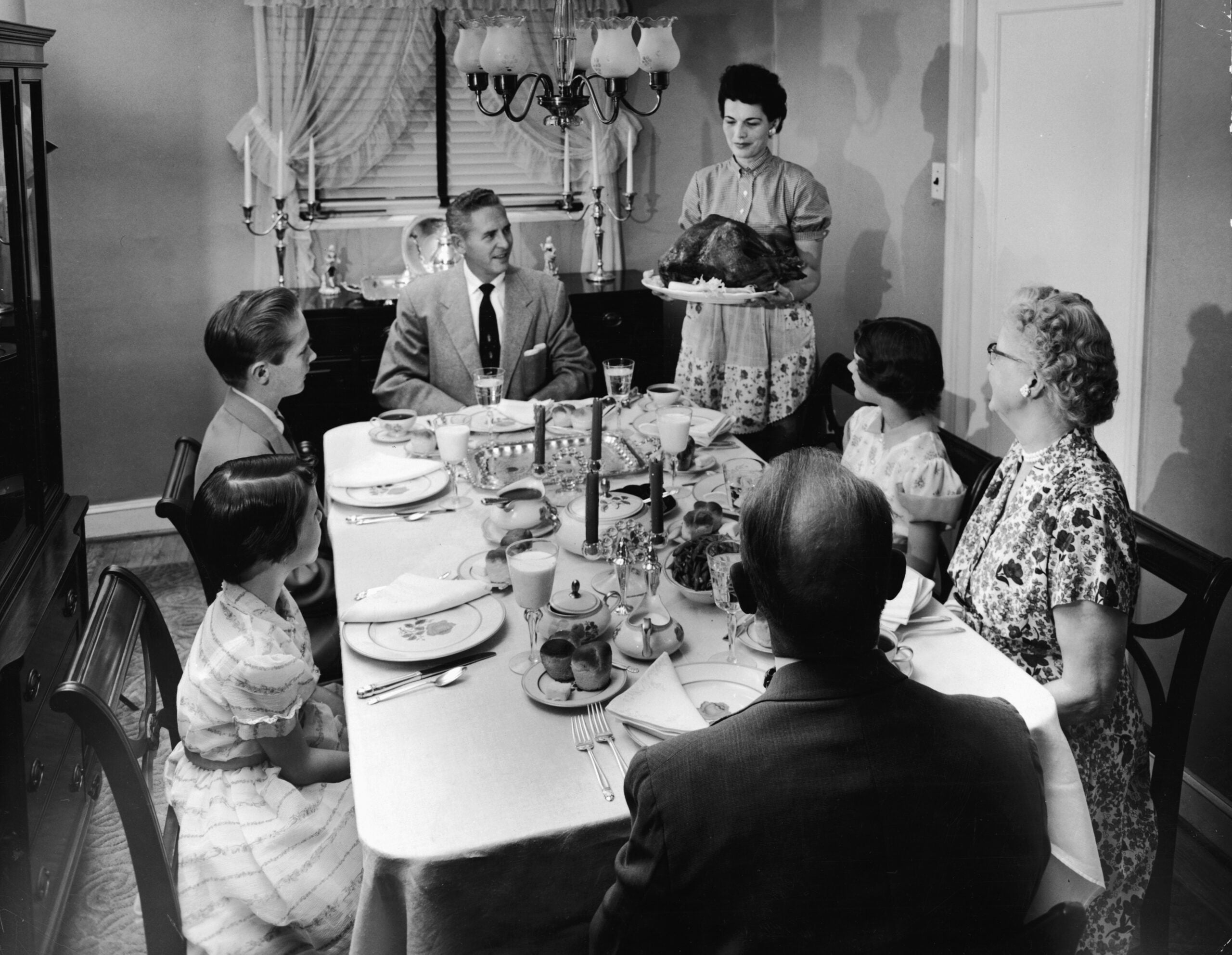 A woman presents a roast turkey to a family of six seated around a dining table, ready for a meal in a 1950s-style dining room.