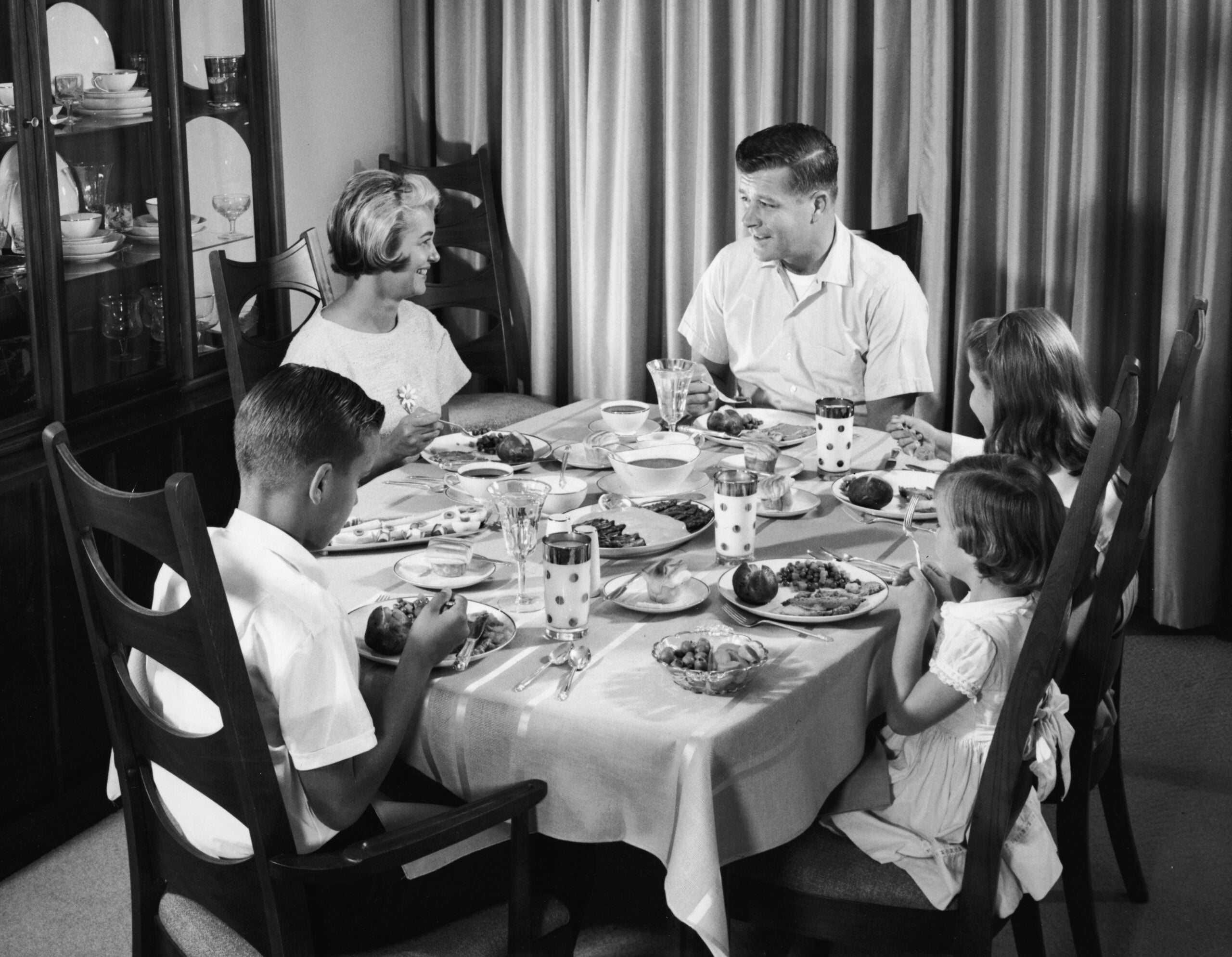 1950s family dinner. A family of five, dressed neatly, sits around a dining table set with plates of food and drinks, smiling and talking together in a warmly lit room with curtains and a cabinet in the background.