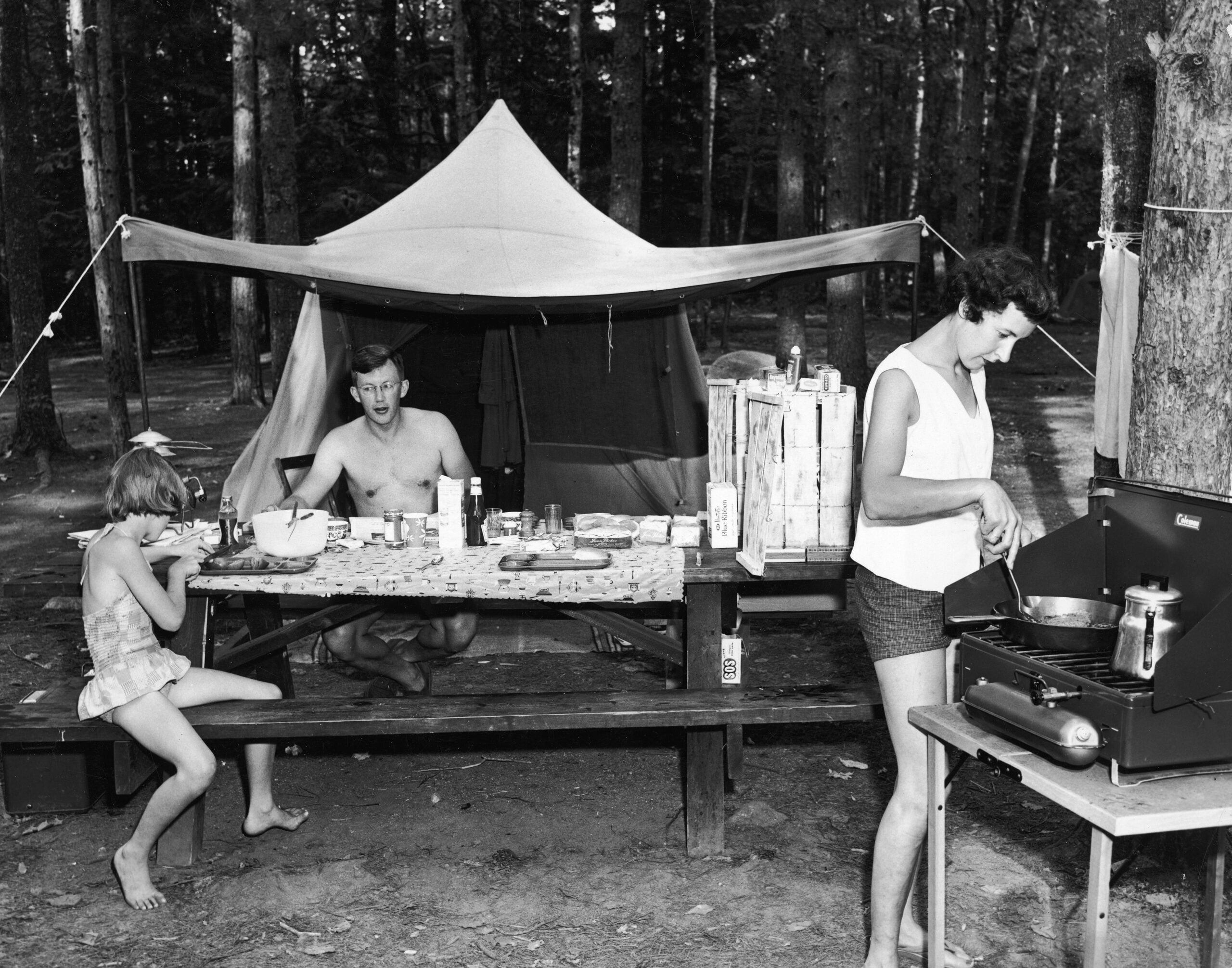 A woman cooks at a camping stove while a man and a child sit at a picnic table in front of a tent, surrounded by trees in a forested campsite.