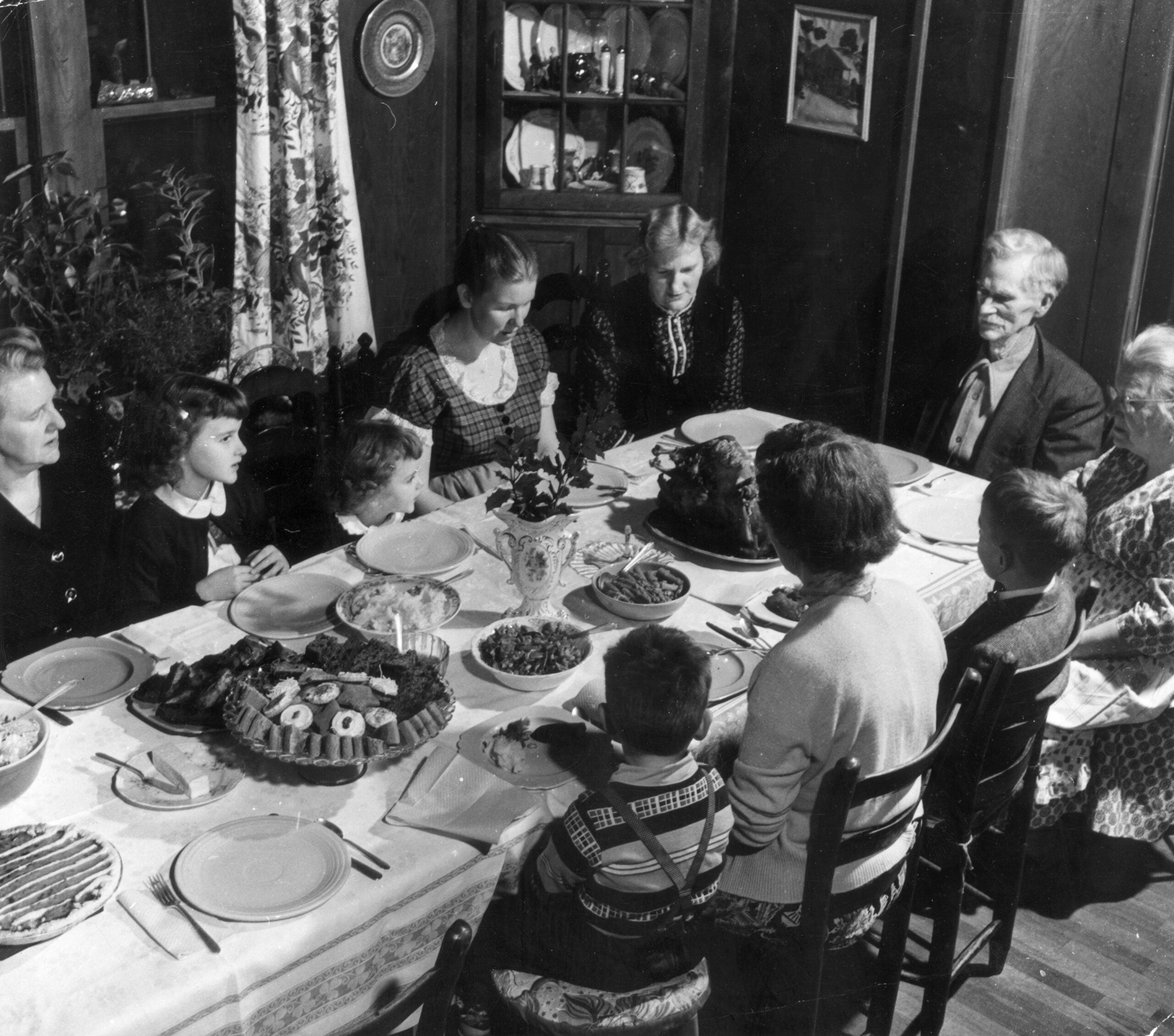 A family of nine, including children and adults, sits around a dining table set with plates and various dishes, sharing a meal in a cozy, vintage home.