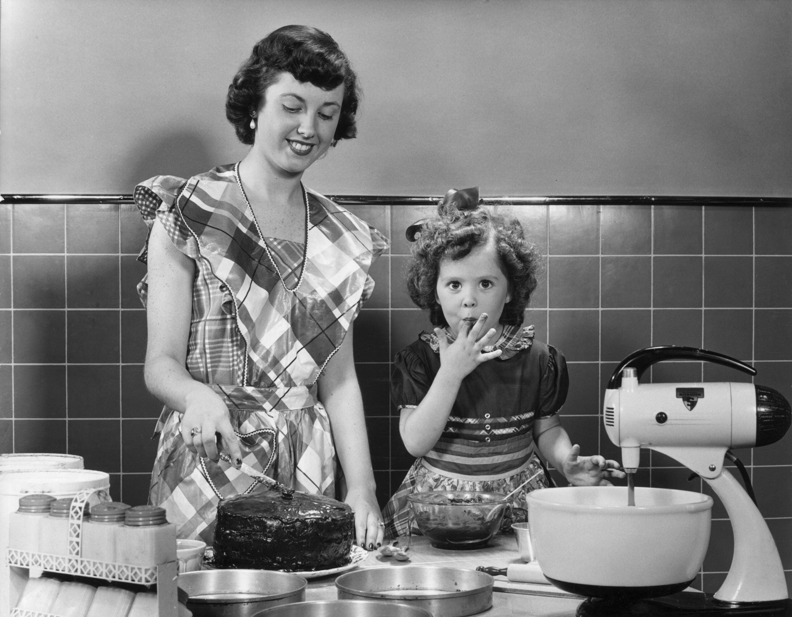 A woman in a plaid dress frosts a cake while a young girl stands beside her, licking batter from her fingers. Mixing bowls and a stand mixer are on the counter. The scene appears vintage, possibly from the 1950s.