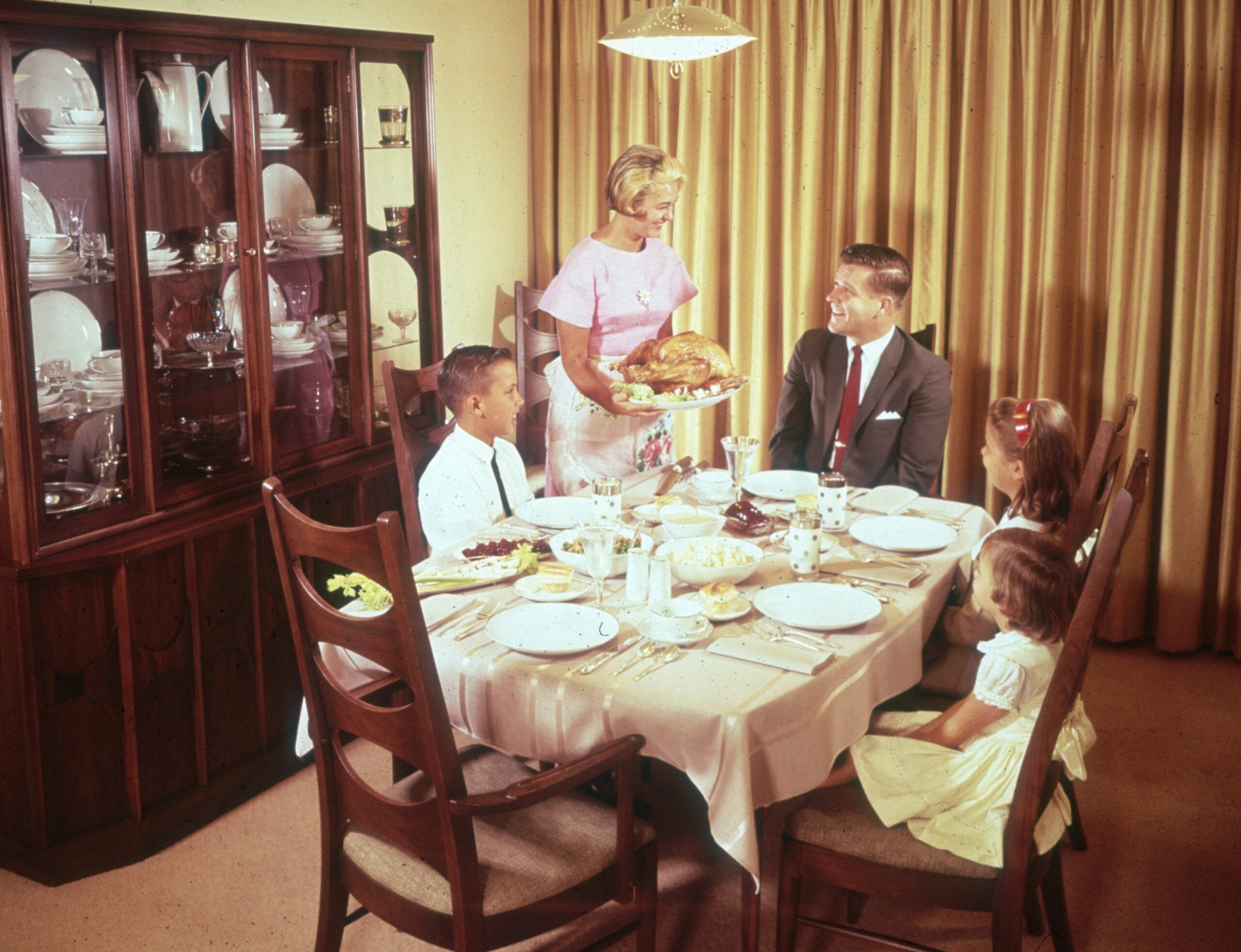 Family dinner in the 1960s. A woman serves a roasted turkey to a man, a boy, and a girl seated at a dining table set with dishes, in a room with a china cabinet and gold curtains.