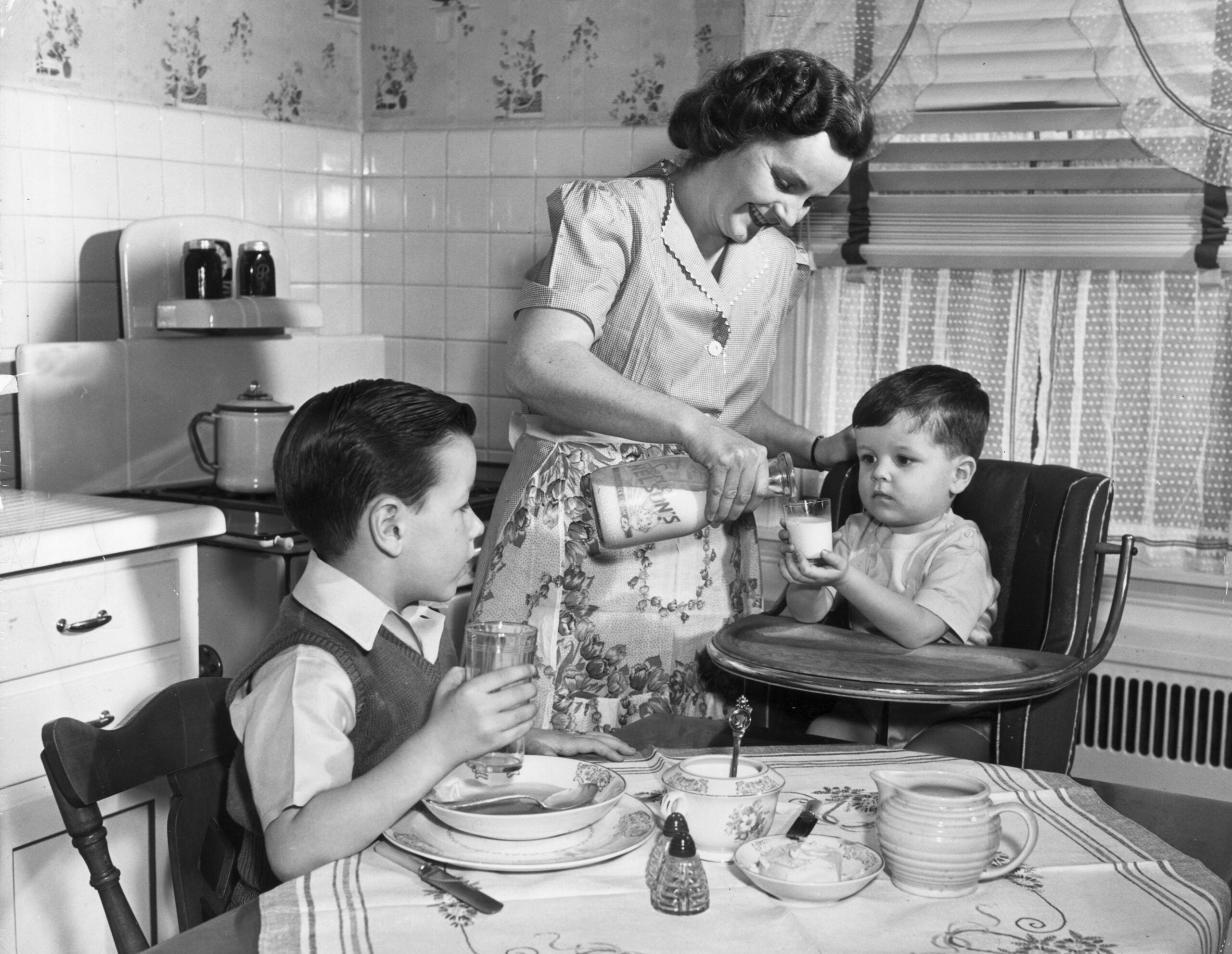 A woman in a 1950s kitchen pours milk for a young boy seated in a high chair while an older boy sits at the table with food and a glass of milk. The scene looks cheerful and domestic.