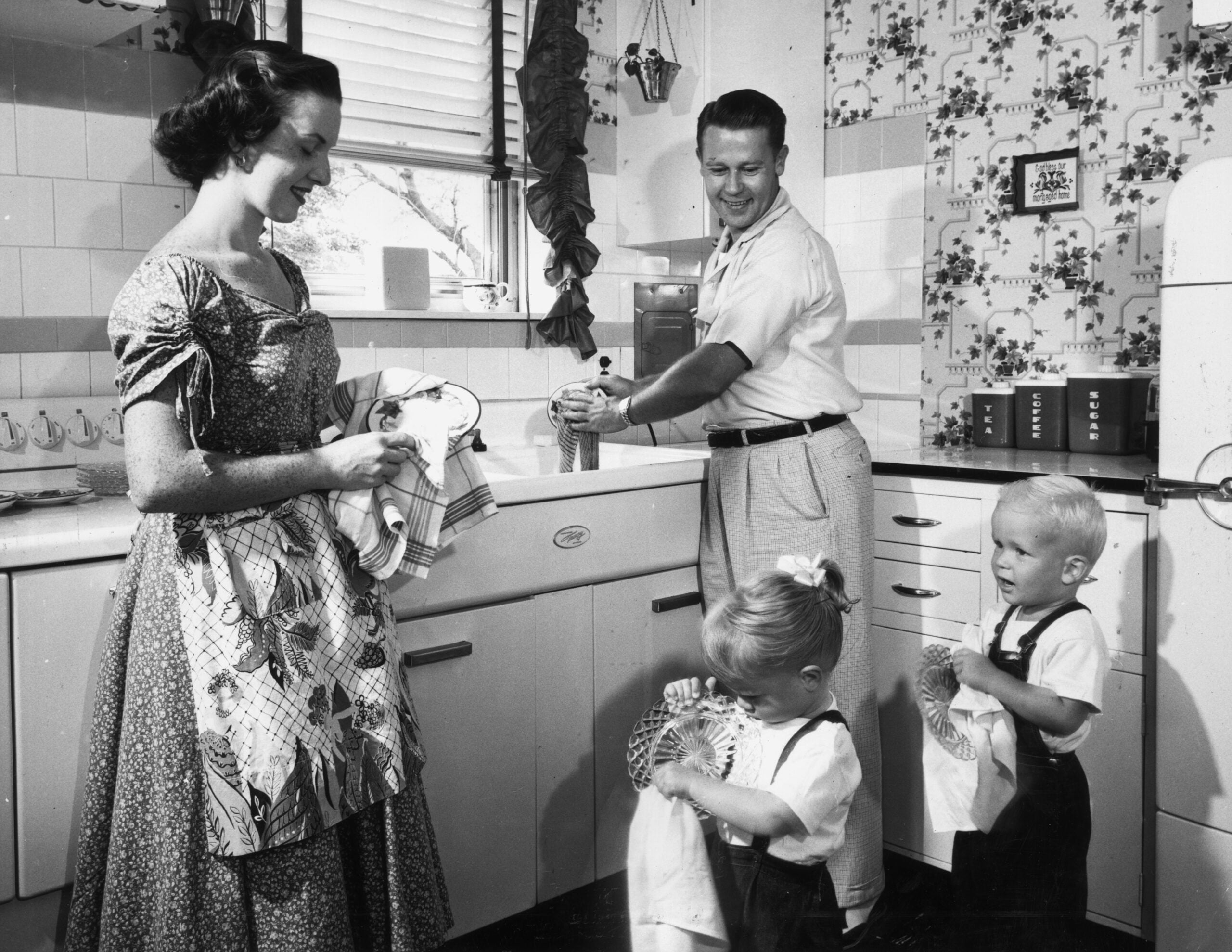 A smiling family of four stands in a 1950s kitchen washing and drying dishes together. The parents and two small children work at the sink, surrounded by vintage decor and patterned wallpaper.