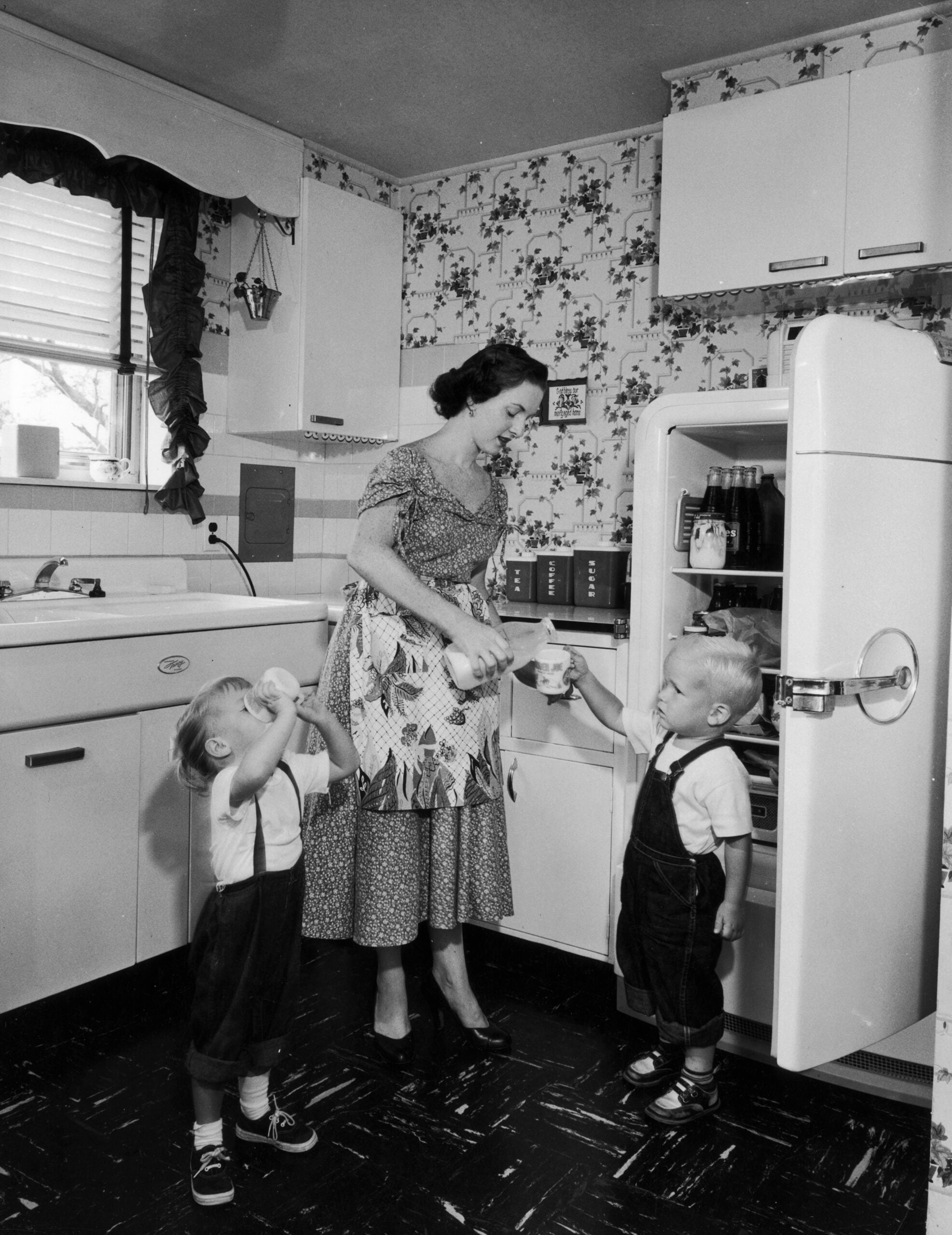 A woman in a patterned dress gives milk to two young children in overalls in a vintage kitchen. The refrigerator door is open and the kids drink from cups.