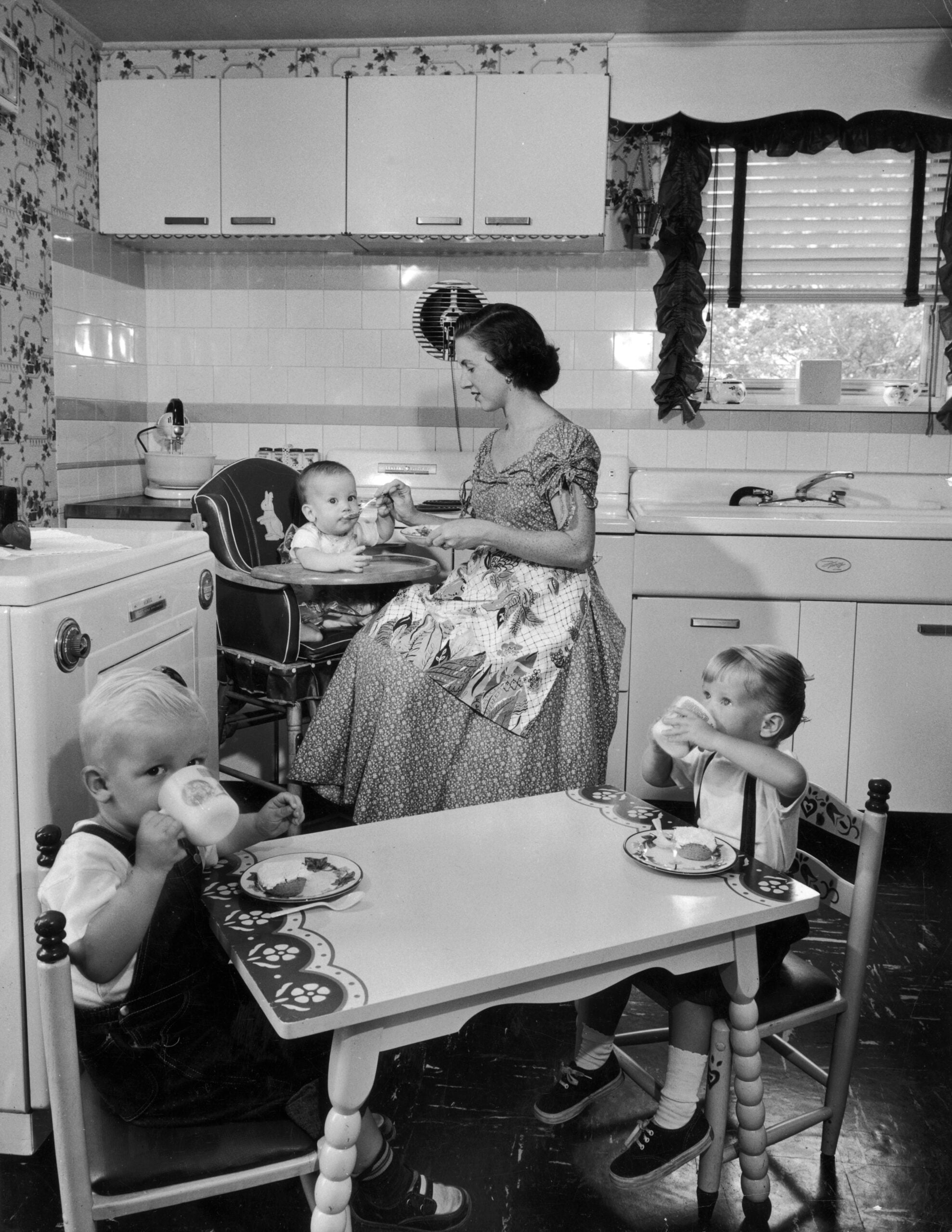 A woman feeds a baby in a high chair while two young children sit at a small table drinking from bottles in a vintage 1950s kitchen.