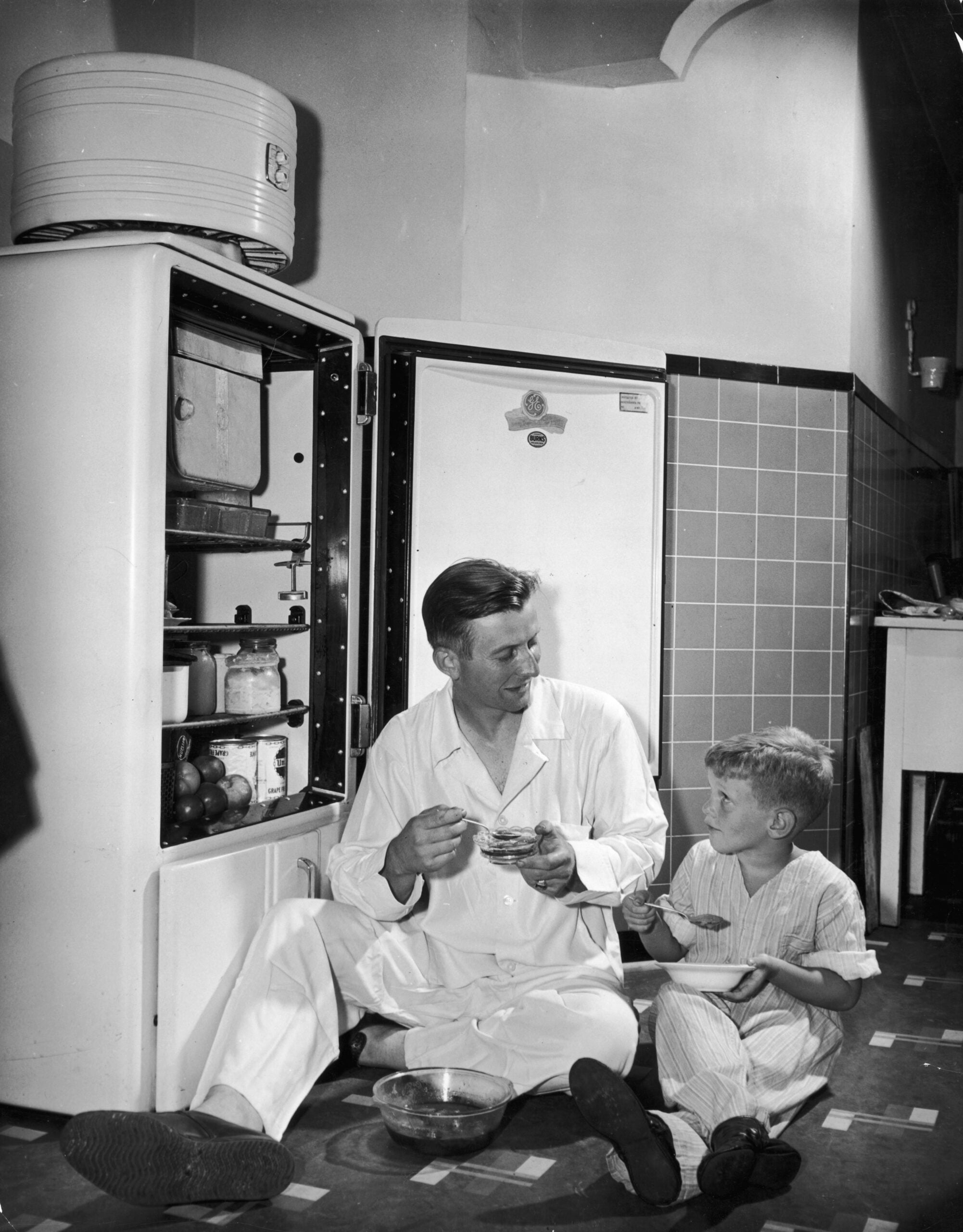 A man and a young boy in pajamas sit on the kitchen floor by an open refrigerator, eating together and smiling. The scene appears to be from a mid-20th century home.