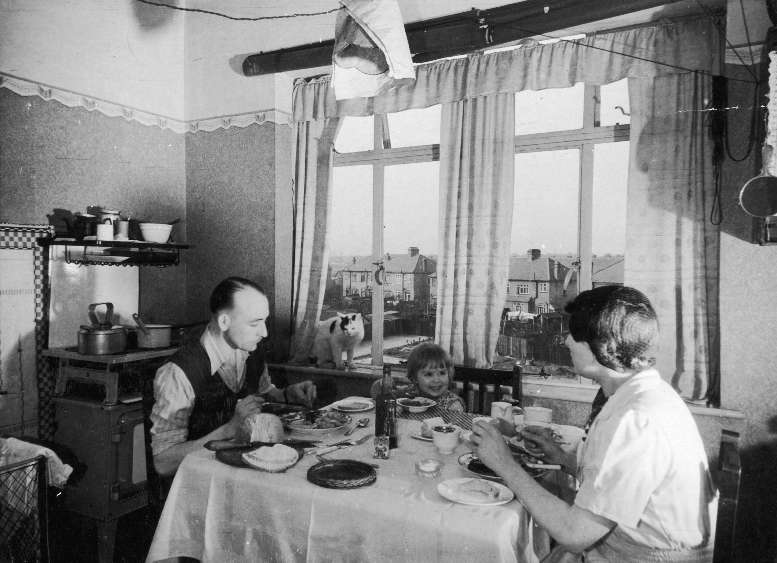 A family of three eats a meal at a kitchen table near a window, with a dog looking in from outside. The room contains shelves, dishes, and a stove, and the window shows houses in the background.