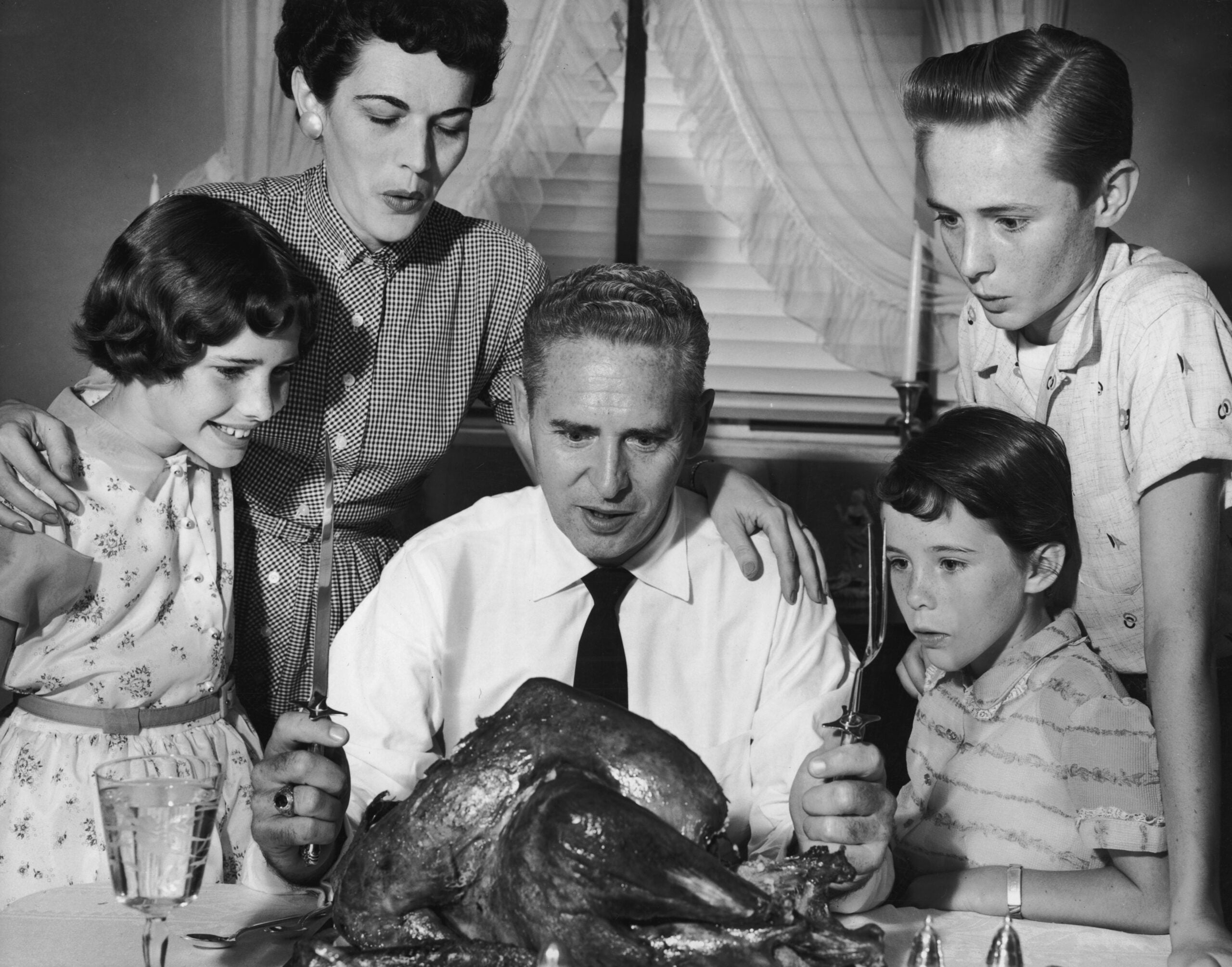 A family of five in vintage clothing gathers around a dining table, eagerly watching as the father prepares to carve a large roasted turkey.