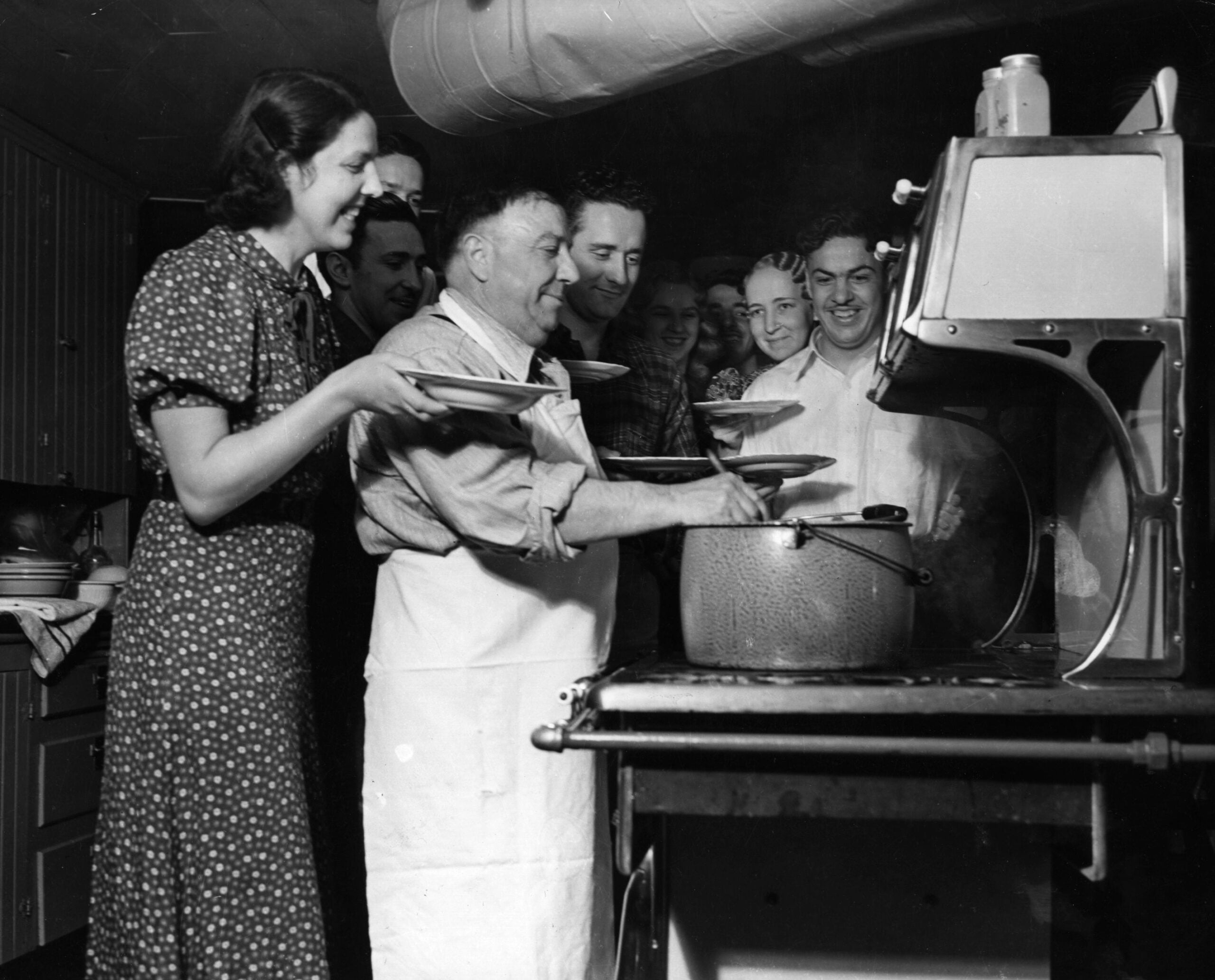 A group of people smiling and waiting in line as a man in an apron serves food from a large pot in a kitchen. A woman in a patterned dress holds a plate at the front of the line.