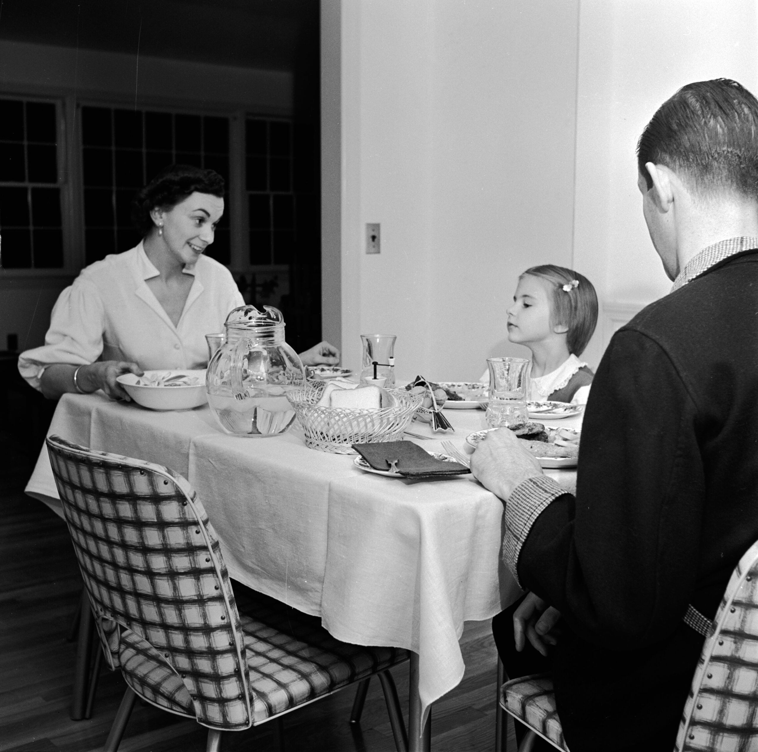 A woman serves food at a dining table to a young girl and a man. The table is set with glassware, a jug, and a basket. The scene appears to be from a mid-20th century home.