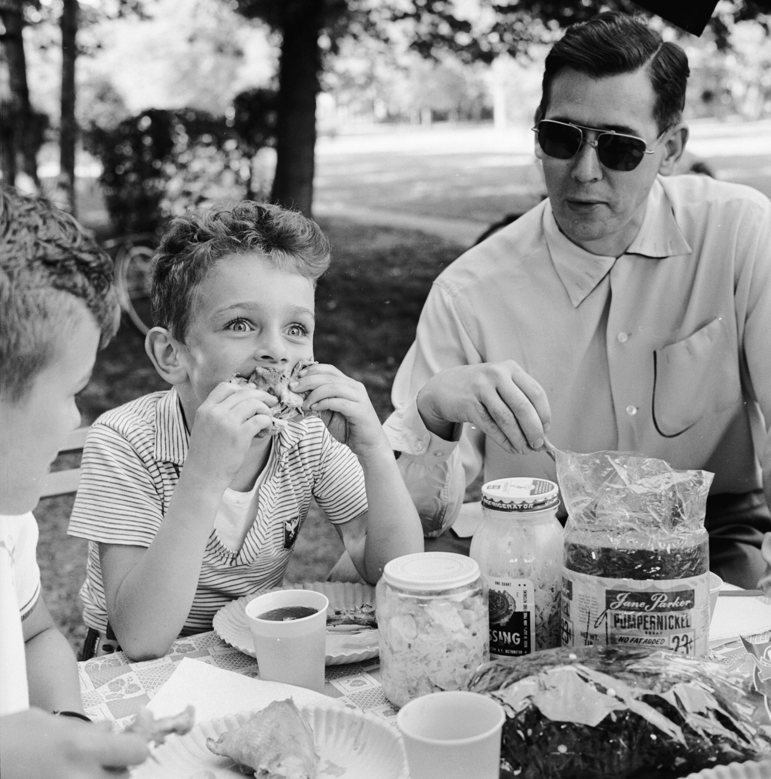 A man and two boys eat at a picnic table outdoors. One boy is biting into a sandwich, while the man wears sunglasses and sits beside him. Various food items, including bread and jars, are on the table.