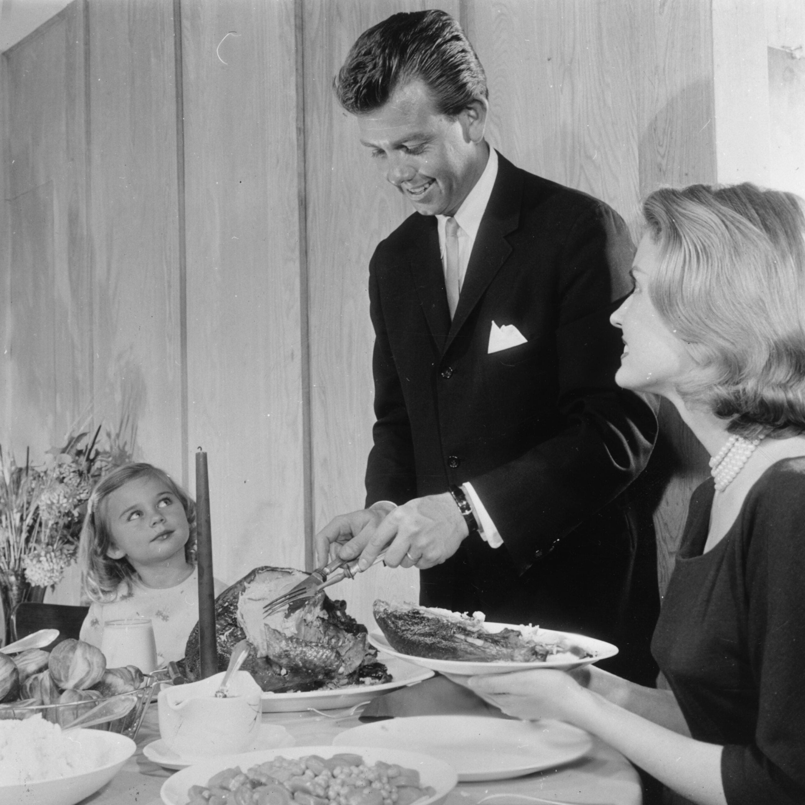 A man in a suit carves a turkey at a dinner table while a woman and a young girl watch. The table is set with food, and the family appears to be enjoying a festive meal together.