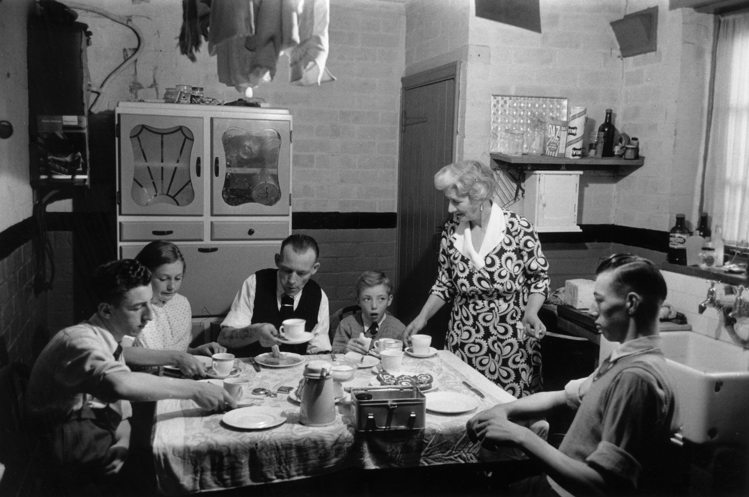 Black-and-white photo of a family of six sitting around a kitchen table, eating a meal together. Dishes, teapot, and cups are on the table. A woman stands, serving or talking, while others are seated.