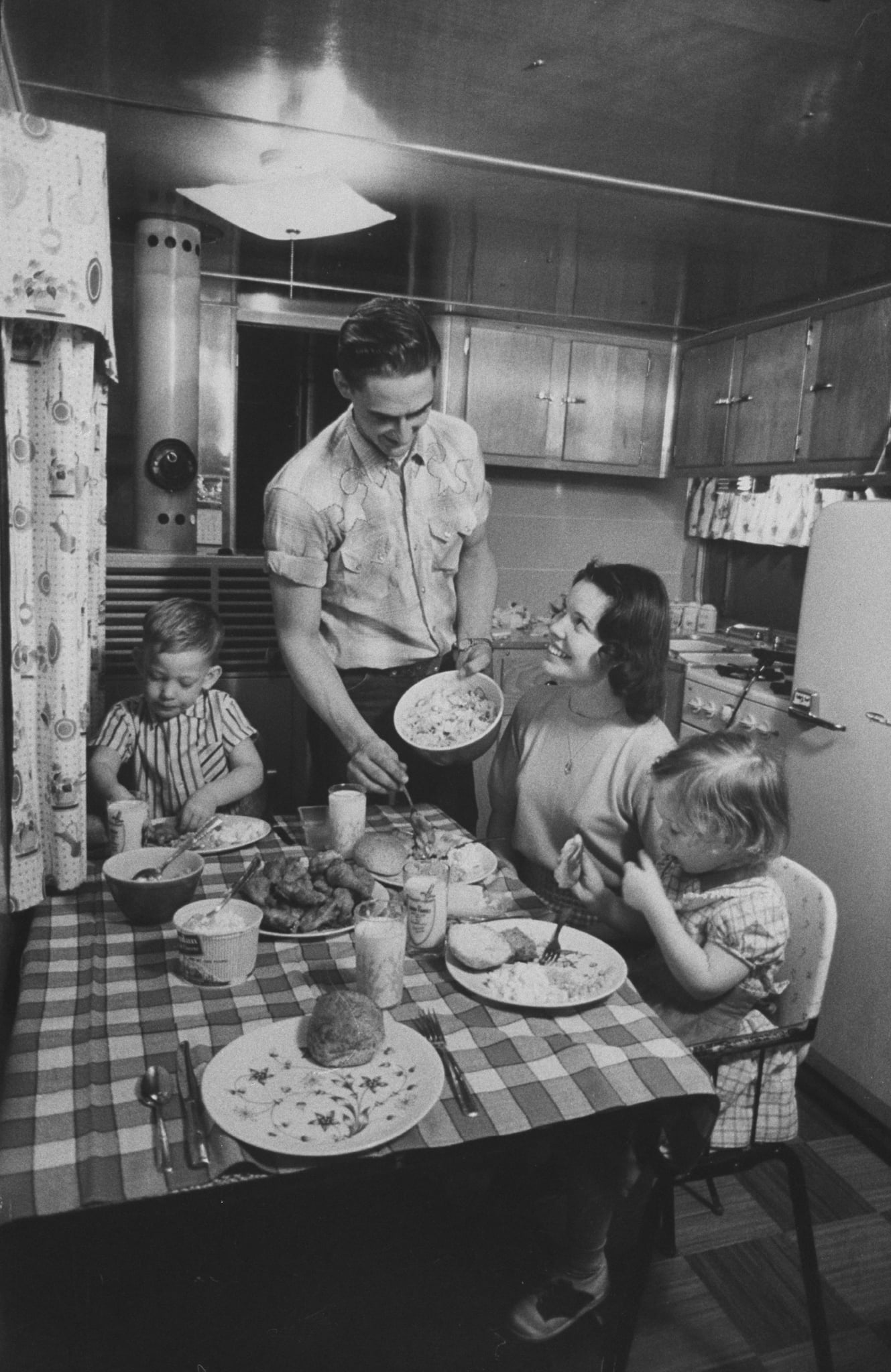 A man serves food to a woman and two young girls at a kitchen table covered with a checkered tablecloth. The kitchen is vintage, with wooden cabinets and a refrigerator in the background.