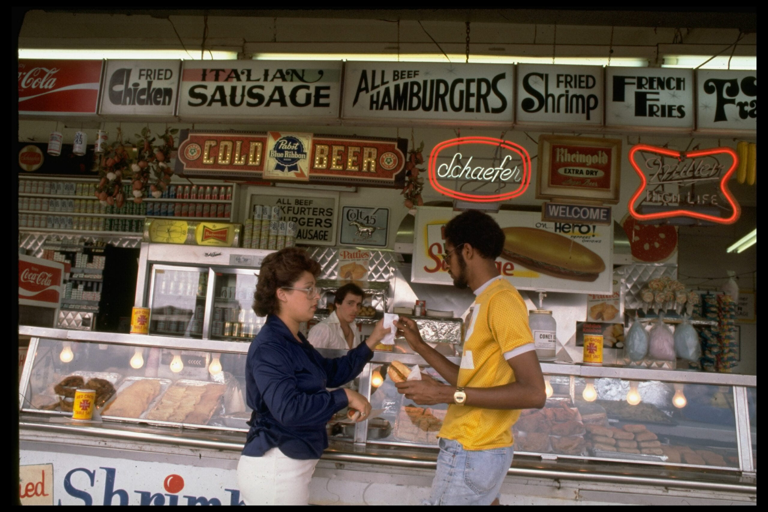 A woman hands food to a man at a busy deli counter, with neon signs above advertising items like fried chicken, sausage, hamburgers, shrimp, cold beer, and soda.