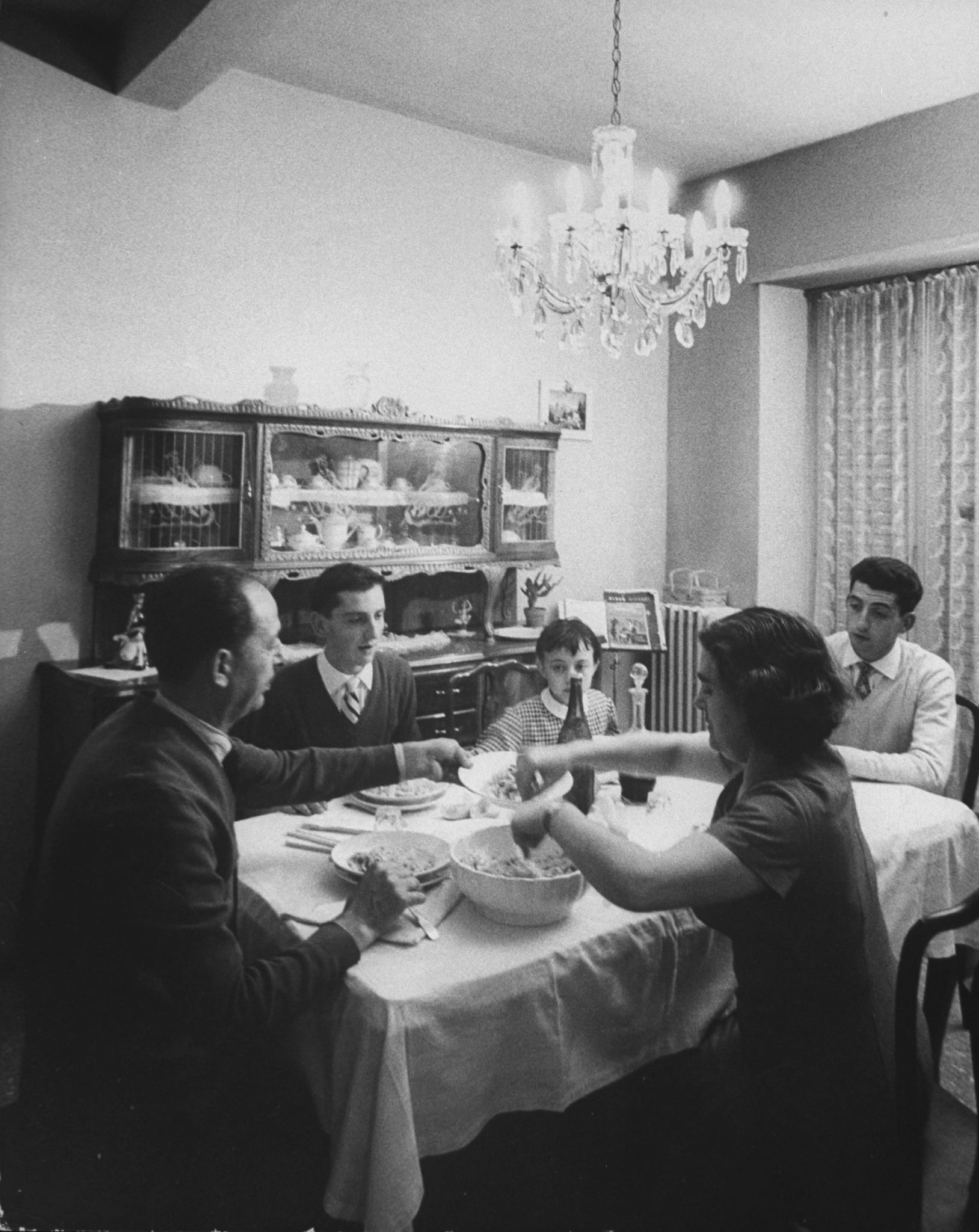 A black-and-white photo of five people sitting around a dining table sharing a meal in a room with a chandelier and a cabinet in the background.
