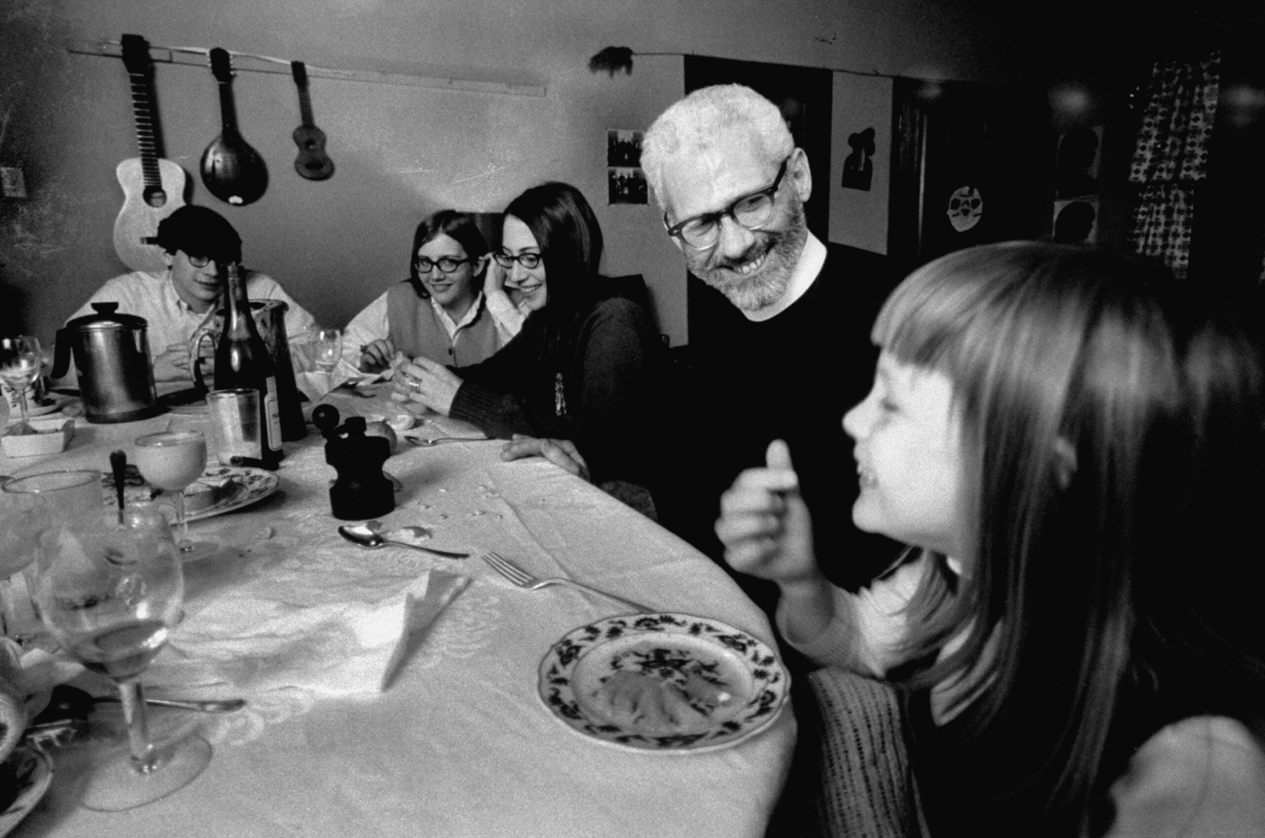 A group of people sit around a dining table, smiling and enjoying a meal together in a cozy room with musical instruments hanging on the wall in the background.