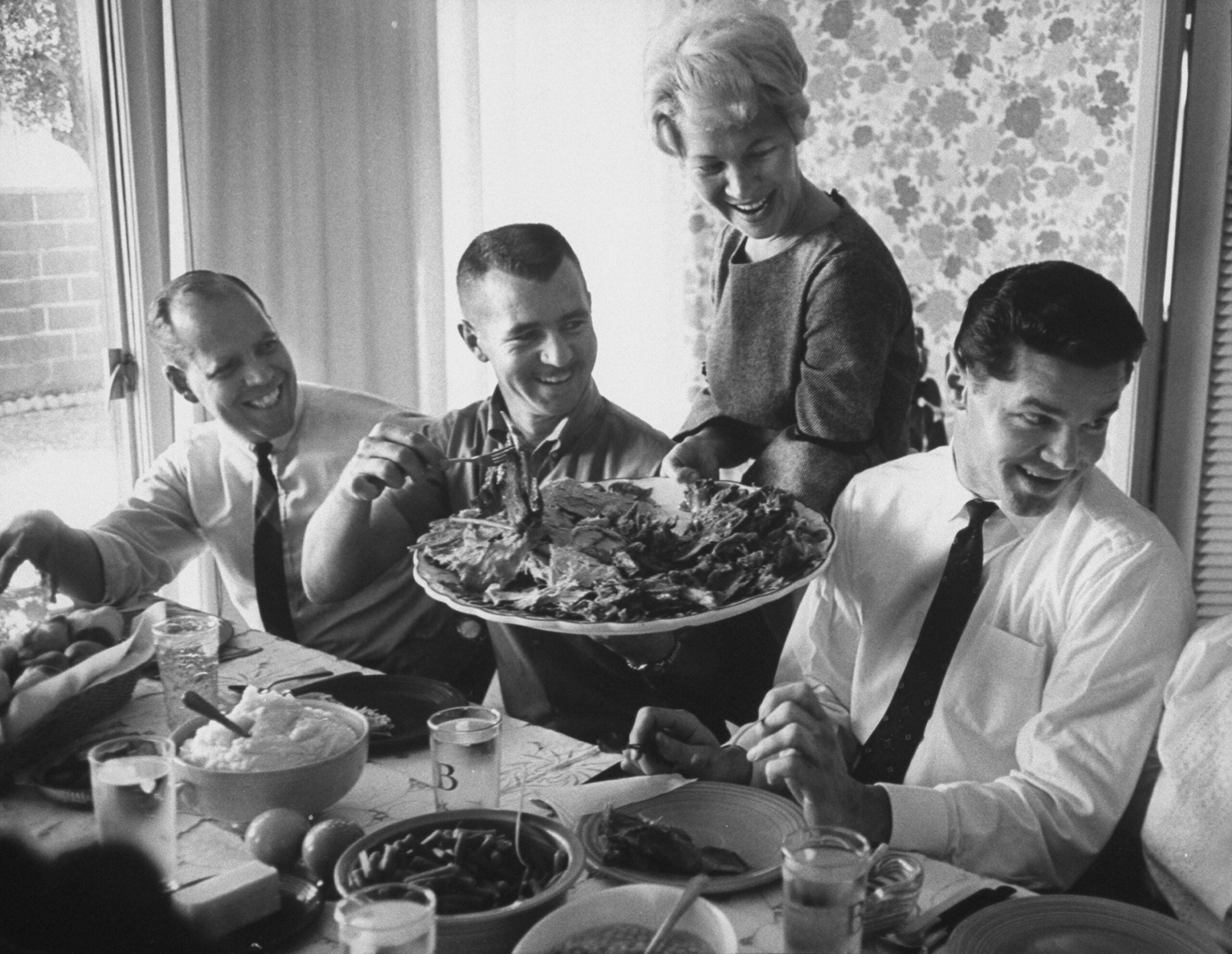 Four people sit at a dining table, smiling and laughing. A woman stands, serving a platter of food. The scene is bright and cheerful, with glasses, bowls, and plates visible on the table.