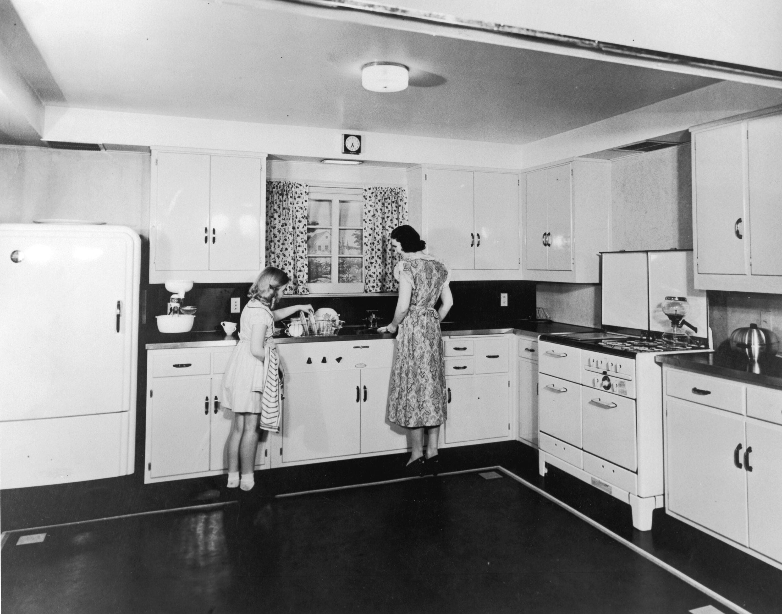 A woman and a girl stand at a kitchen sink, washing dishes. The kitchen has vintage appliances, white cabinets, and a window with patterned curtains.