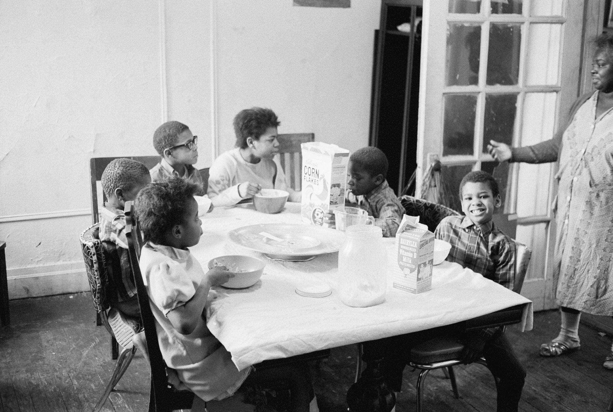 Six children sit around a dining table eating breakfast cereal. A woman stands nearby gesturing with her hand. The table has bowls, cereal boxes, and a pitcher. The room appears homey and modest.
