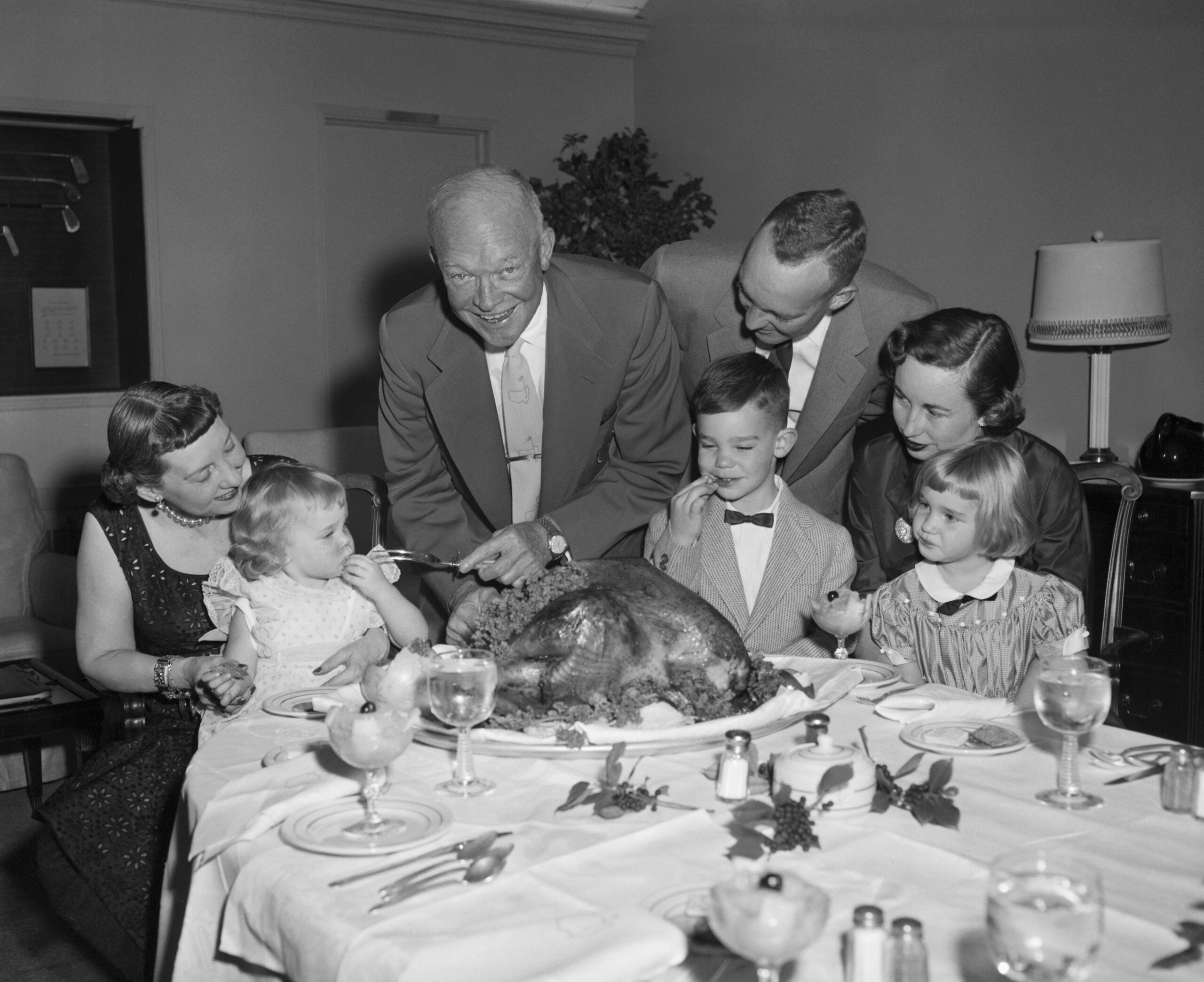 An older man carves a turkey at a dinner table surrounded by smiling adults and children, suggesting a family gathering or holiday meal.