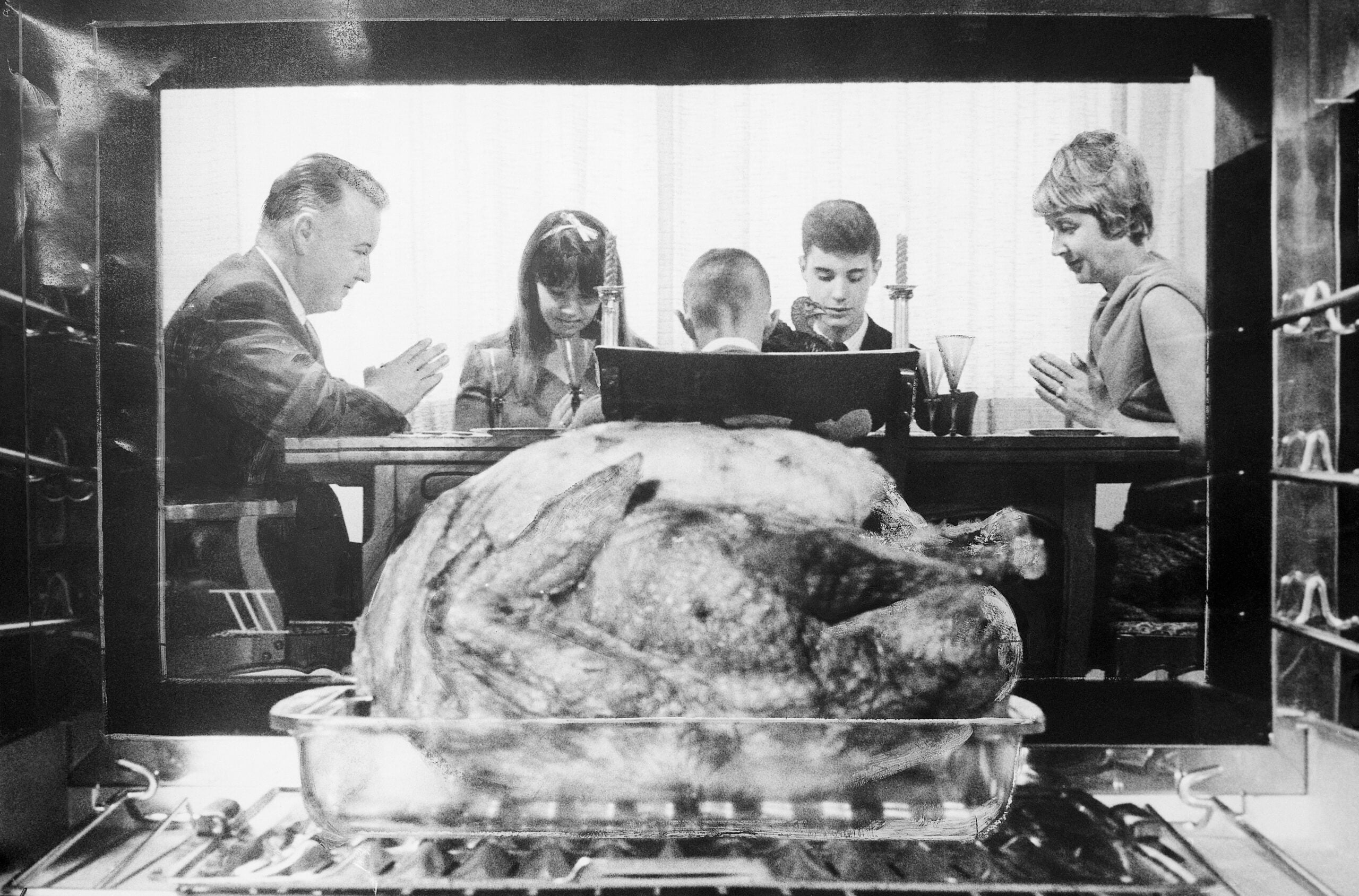 A family sits around a table, heads bowed in prayer, as seen through an oven with a large roasted turkey in the foreground.
