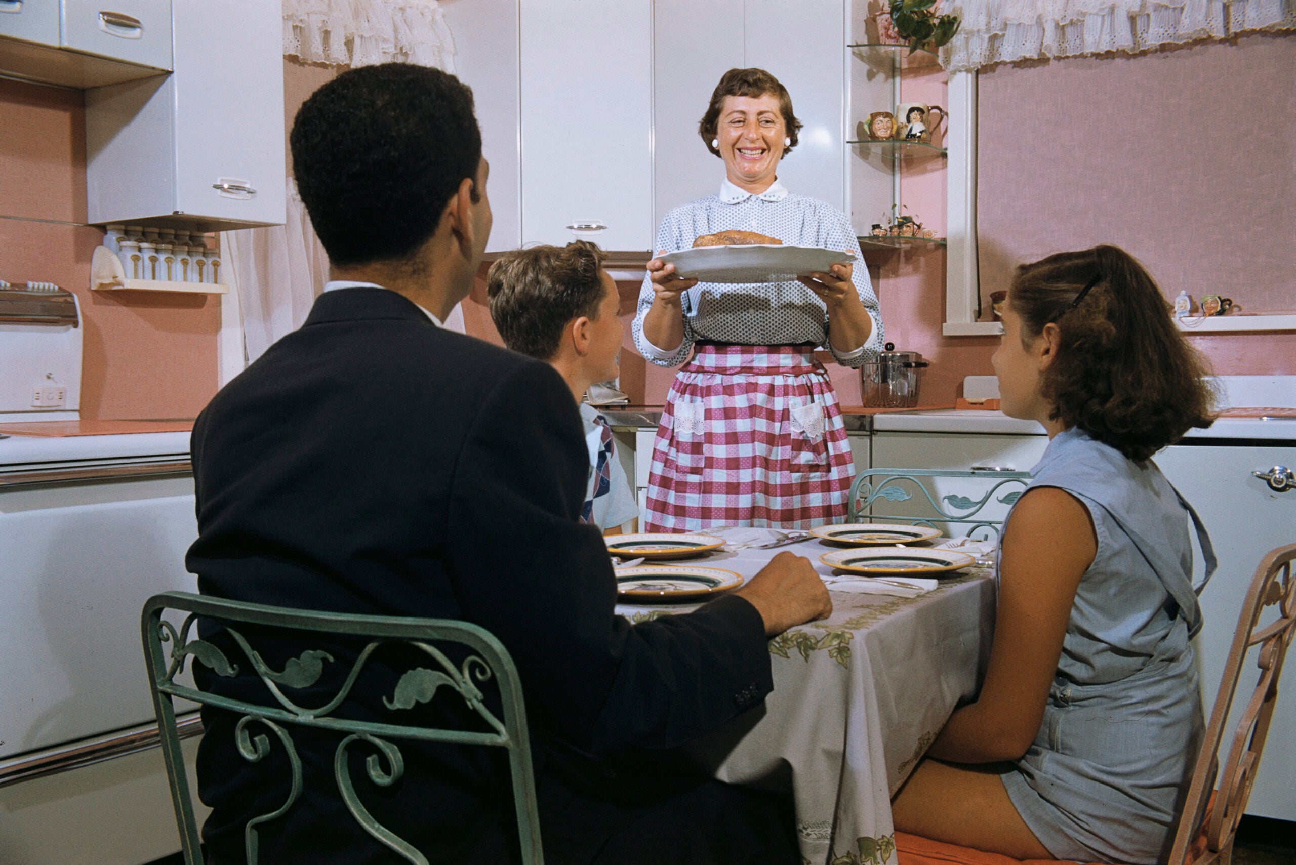 A woman in an apron smiles as she serves a pie to three people seated at a kitchen table in a retro-style kitchen.