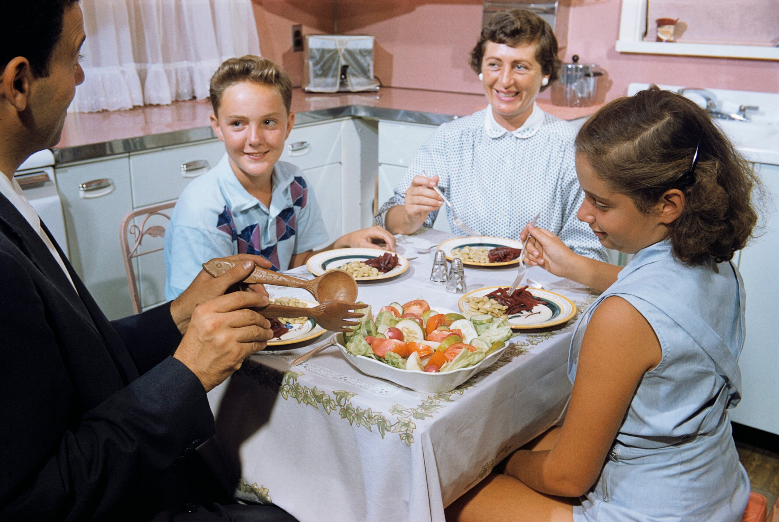 A family of four sits around a kitchen table, smiling, eating dinner together. There is a large salad bowl in the center and plates of food in front of each person. The kitchen has light-colored cabinets and curtains.