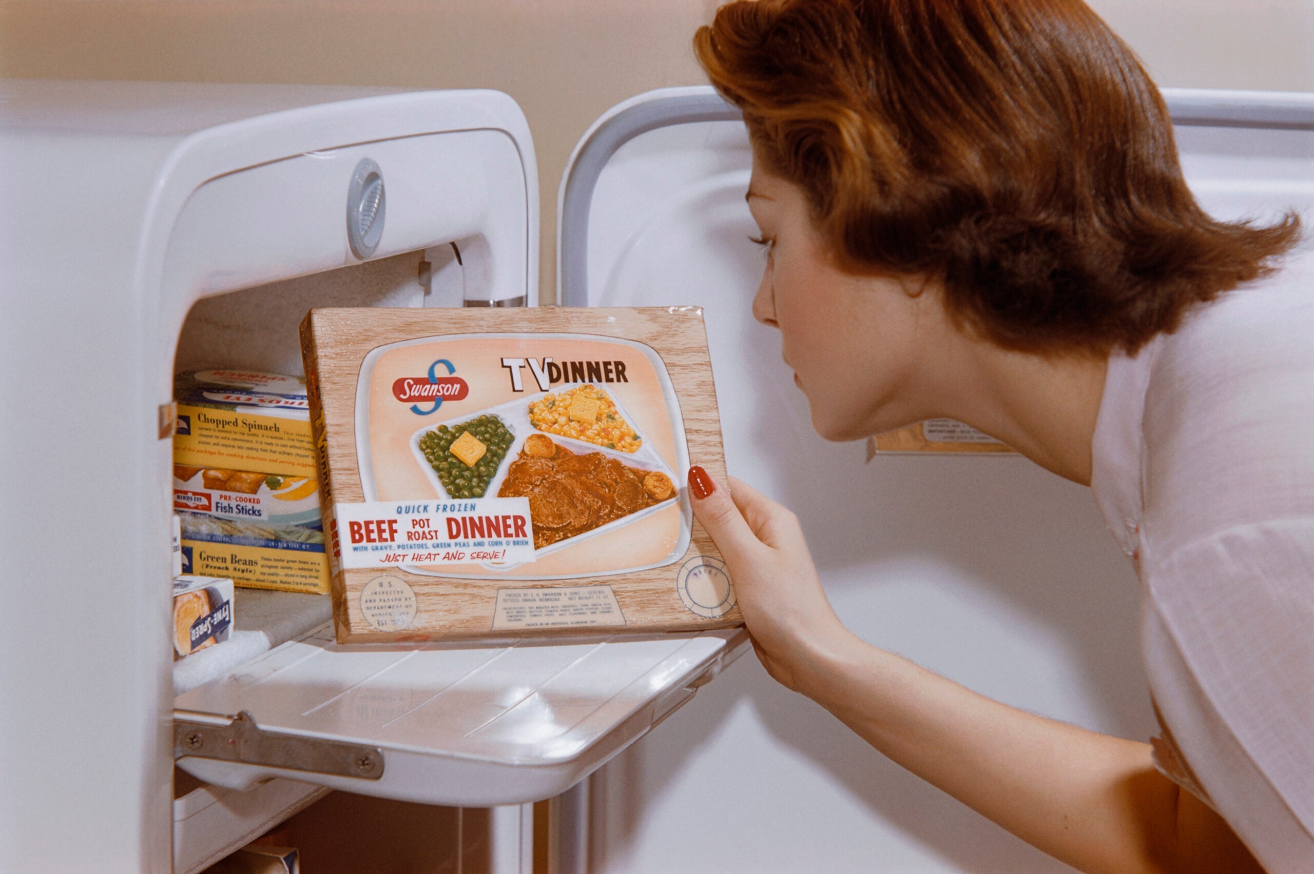 Popular foods in the 60s and 70s. A woman takes a Swanson TV Dinner out of a freezer. Frozen food boxes, including spinach, are visible inside. The TV Dinner box shows beef, mashed potatoes, corn, and a dessert.