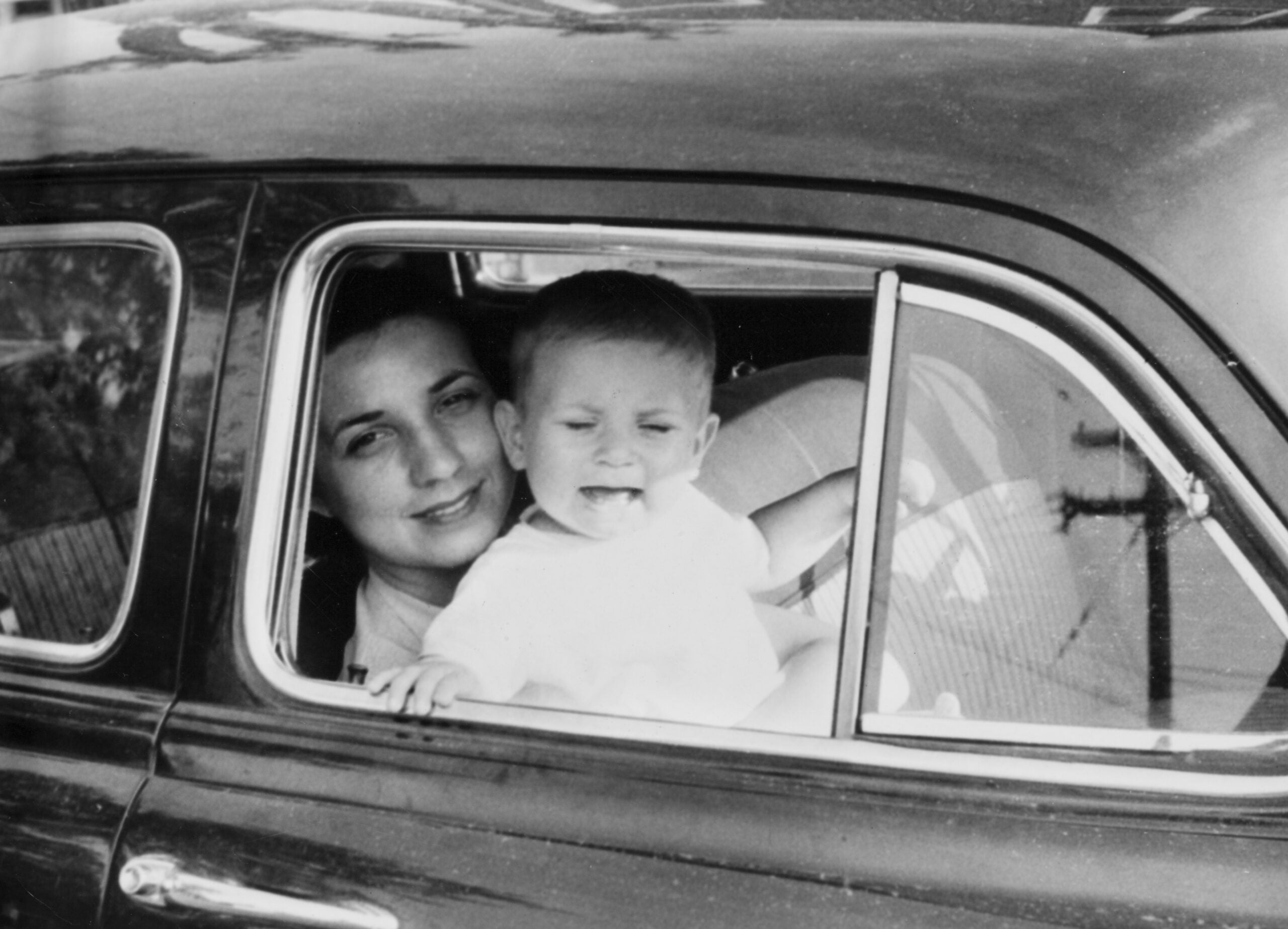 A woman smiles while holding a baby who is sticking out his tongue, both looking out of the open window of a vintage car, capturing the joy and spontaneity of family road trips.