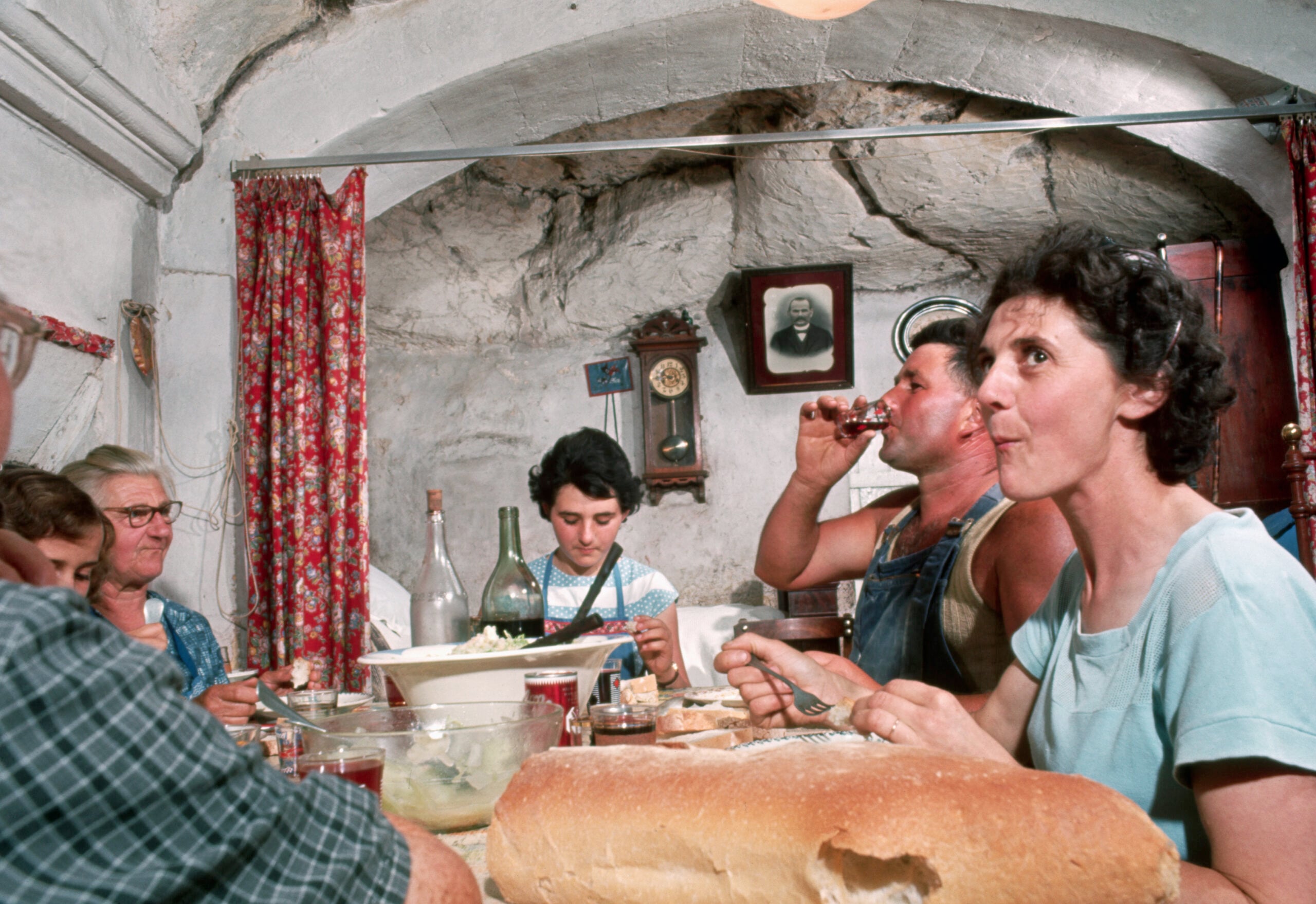 Six people sit around a table in a stone-walled room, sharing a meal with bread, wine, and salad. A clock and a portrait hang on the wall, and red curtains frame a window in the background.