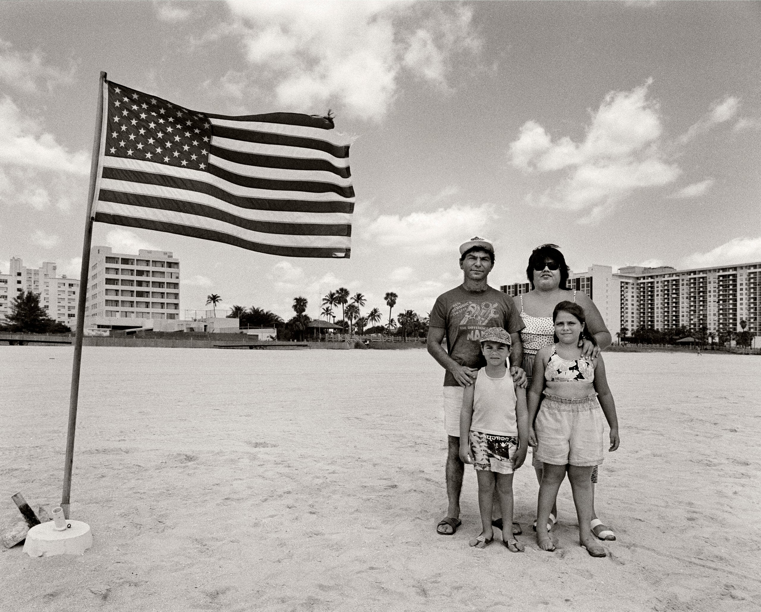 A family of four stands on a sandy beach near a large American flag, with tall buildings, palm trees, and a partly cloudy sky in the background.