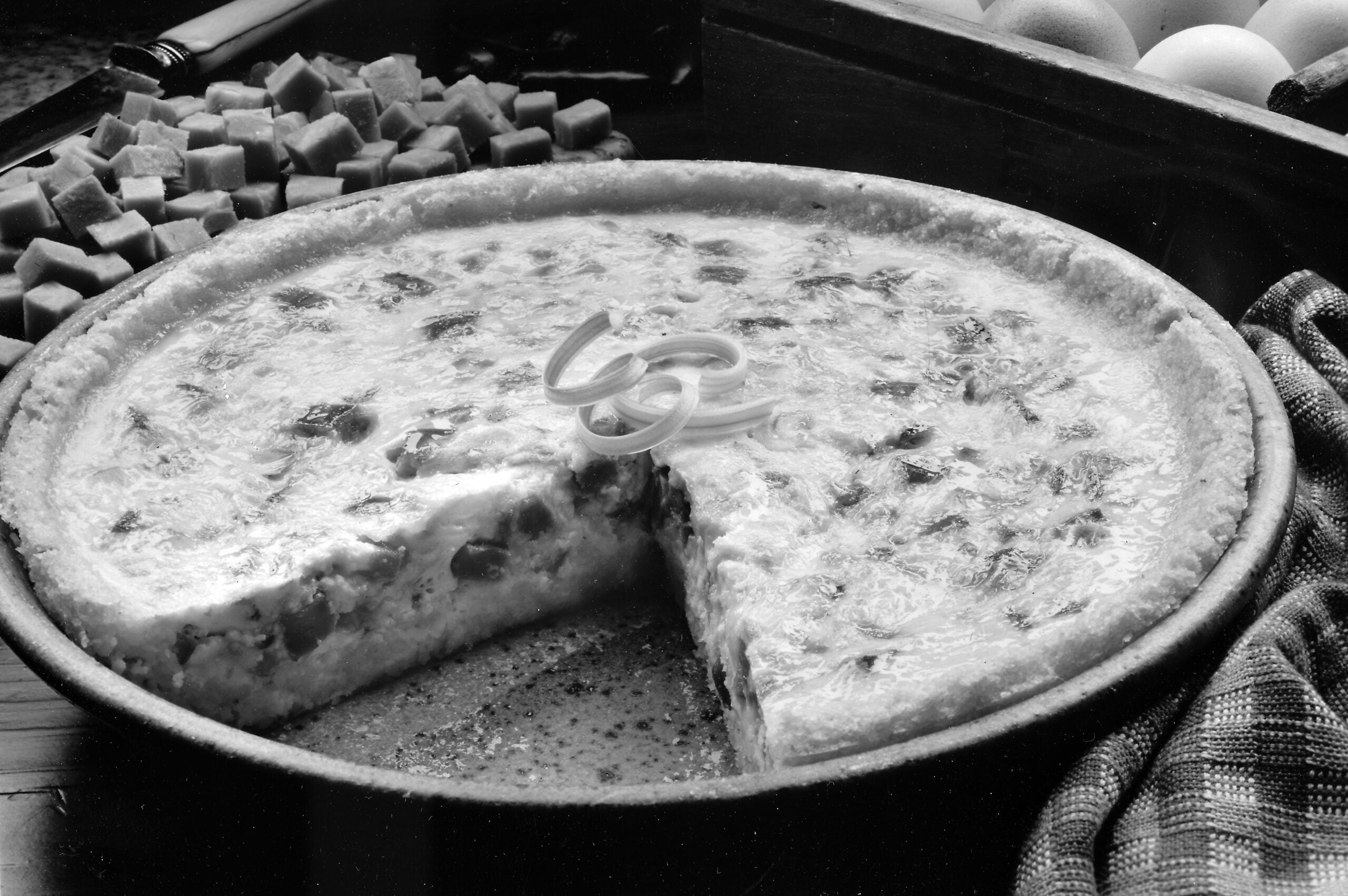A quiche with a slice missing sits on a table, garnished with onion rings. Diced ham, whole eggs, and a plaid cloth are nearby.