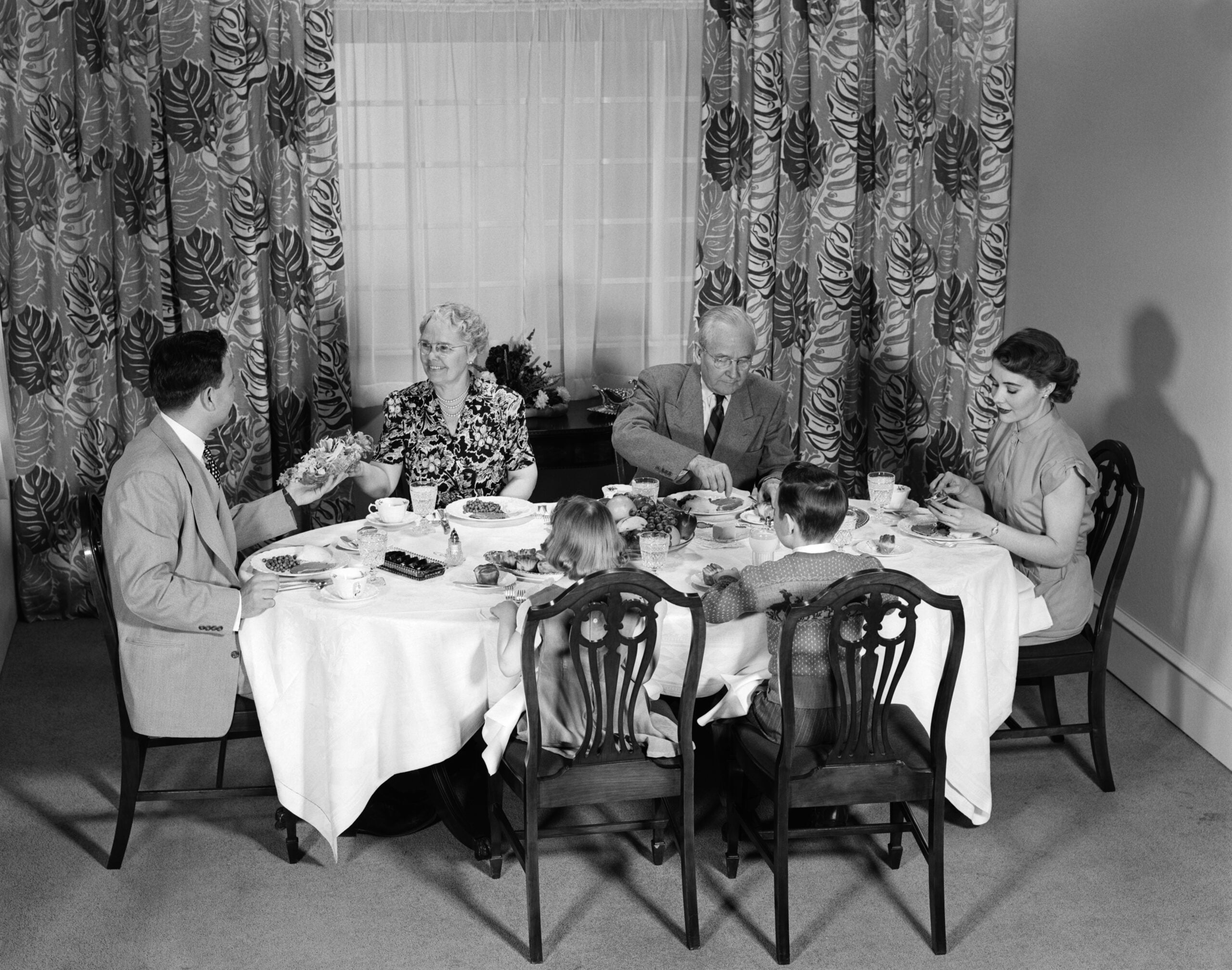 A black-and-white photo of six people—three adults and three children—sitting around a round dining table, sharing a meal in a room with patterned curtains and a window in the background.