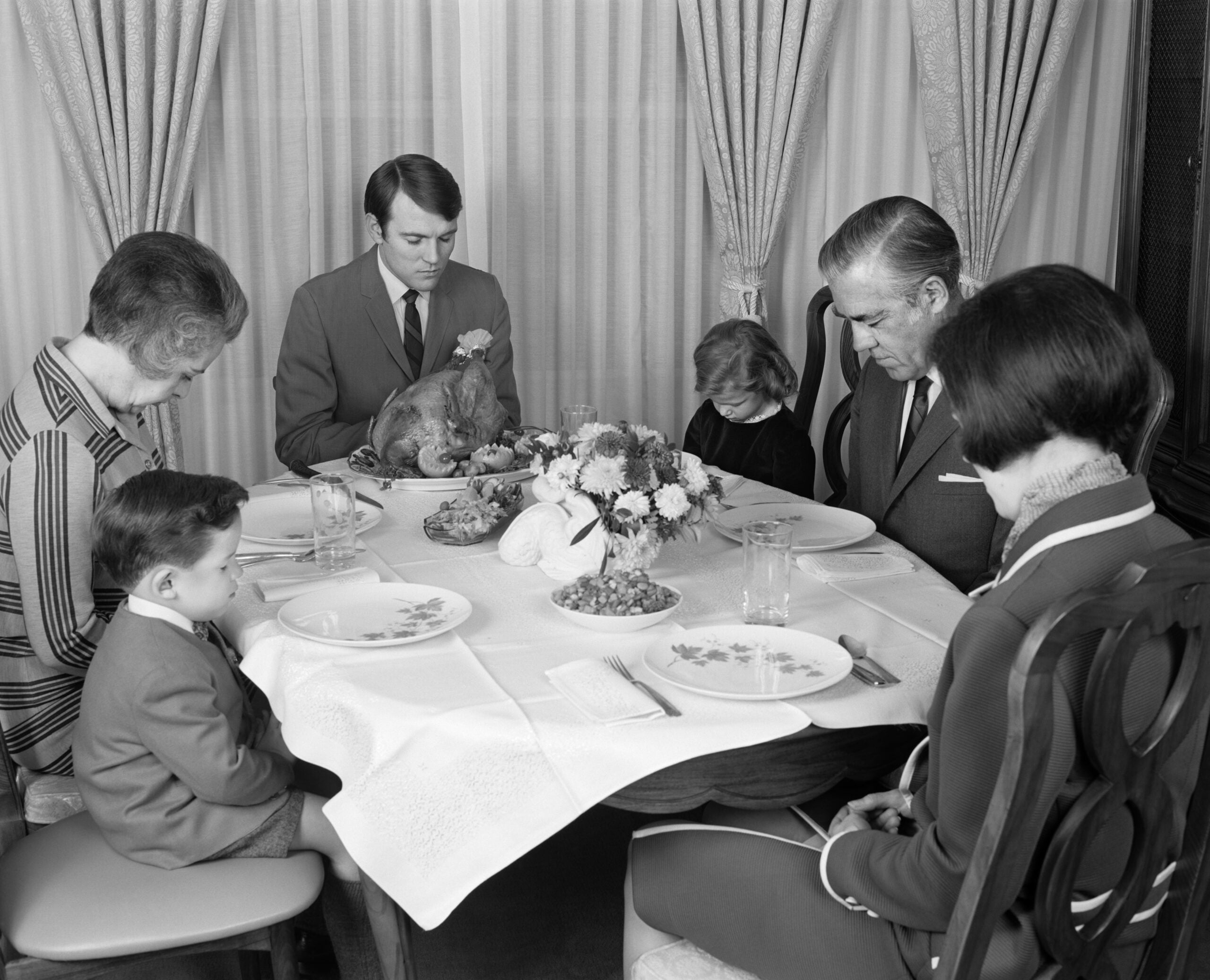 A family of six, including two children, sits around a table with a turkey centerpiece, heads bowed in prayer before a meal. The photo is black and white and set in a formal dining room.