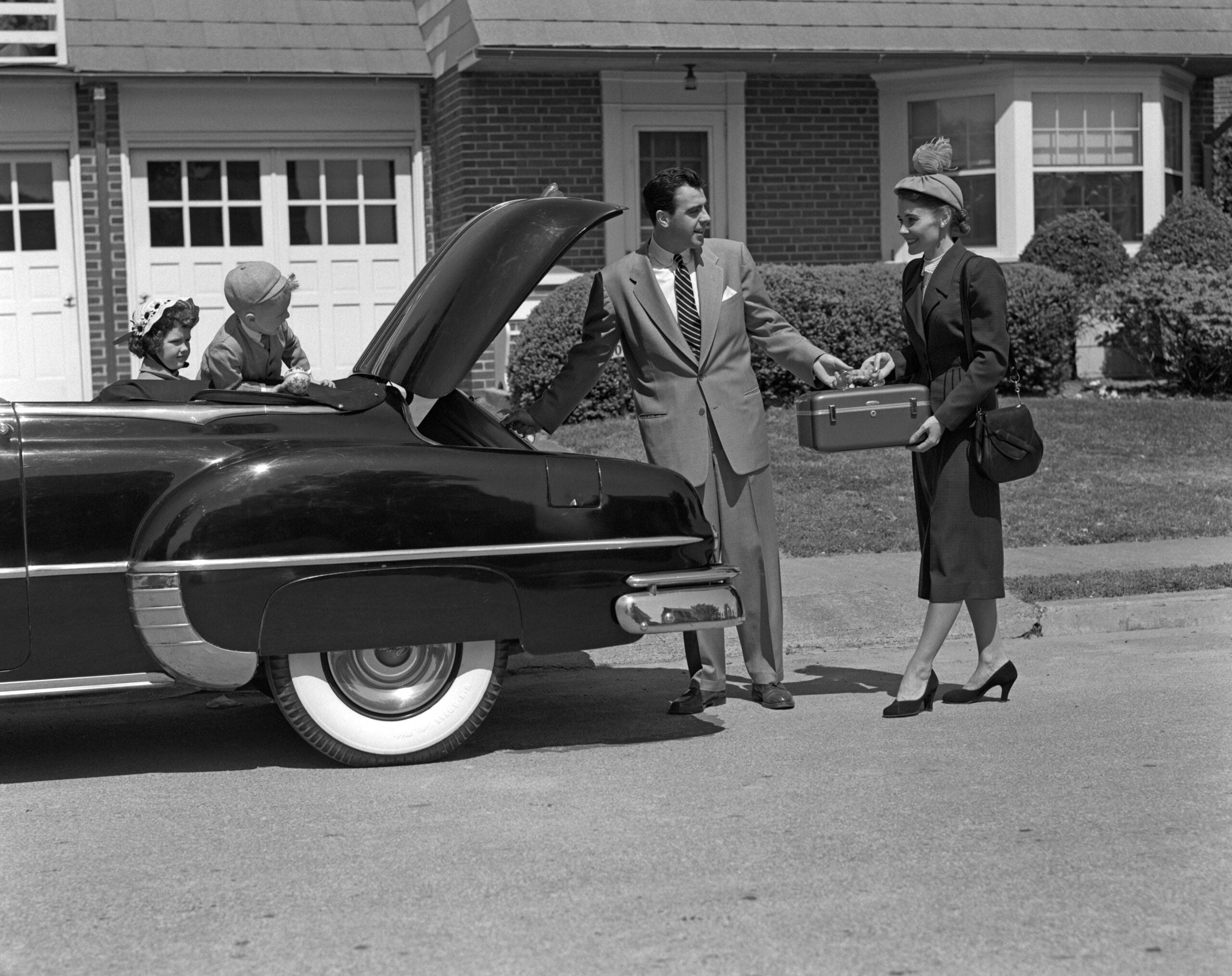 A man in a suit helps a woman in a hat with her suitcase beside a classic car, while two children sit in the back seat—capturing the excitement and togetherness of family road trips—in front of a suburban house.