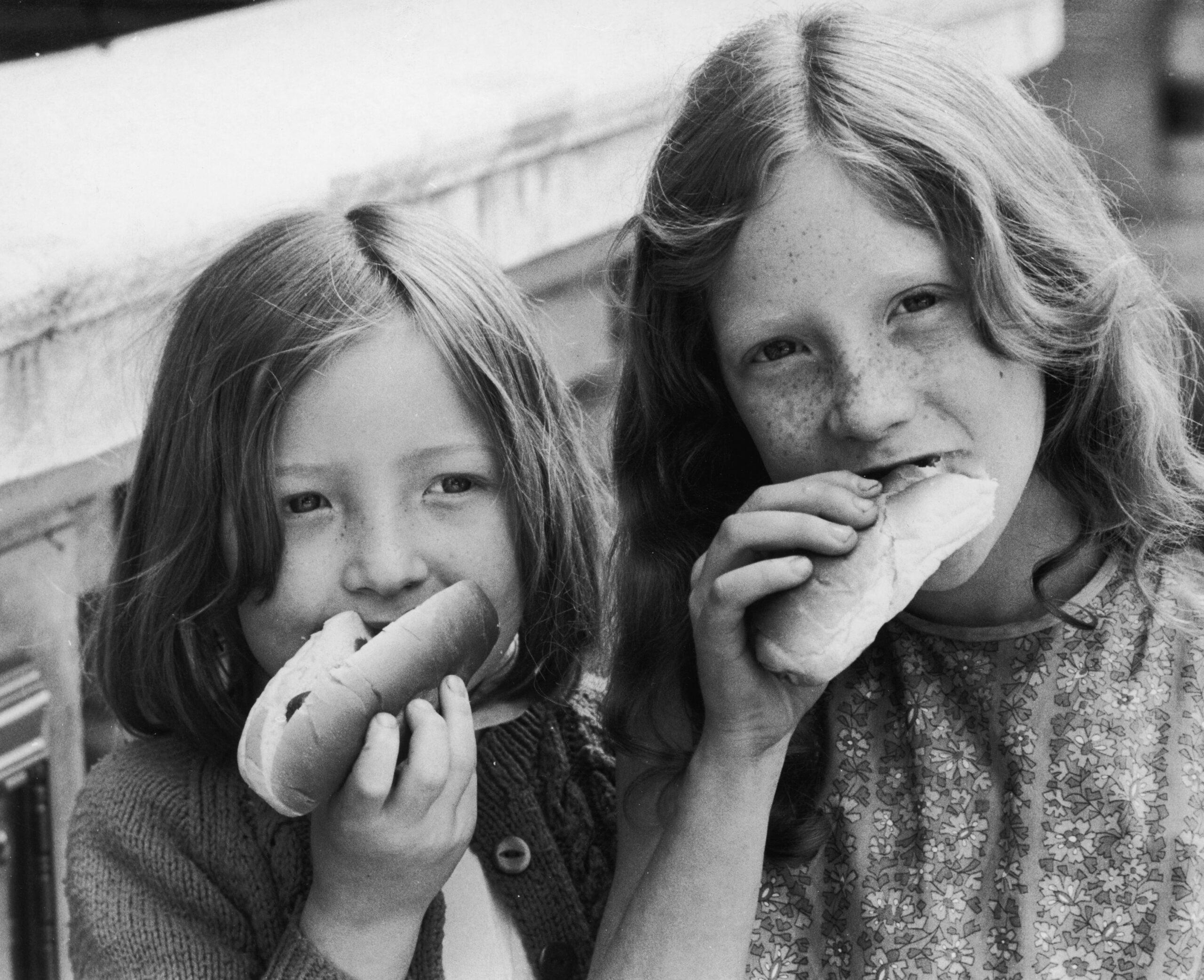 Two young girls with long hair eat hot dogs, smiling at the camera. One girl wears a patterned dress, the other a cardigan. They appear to be outside, possibly at a picnic or gathering.