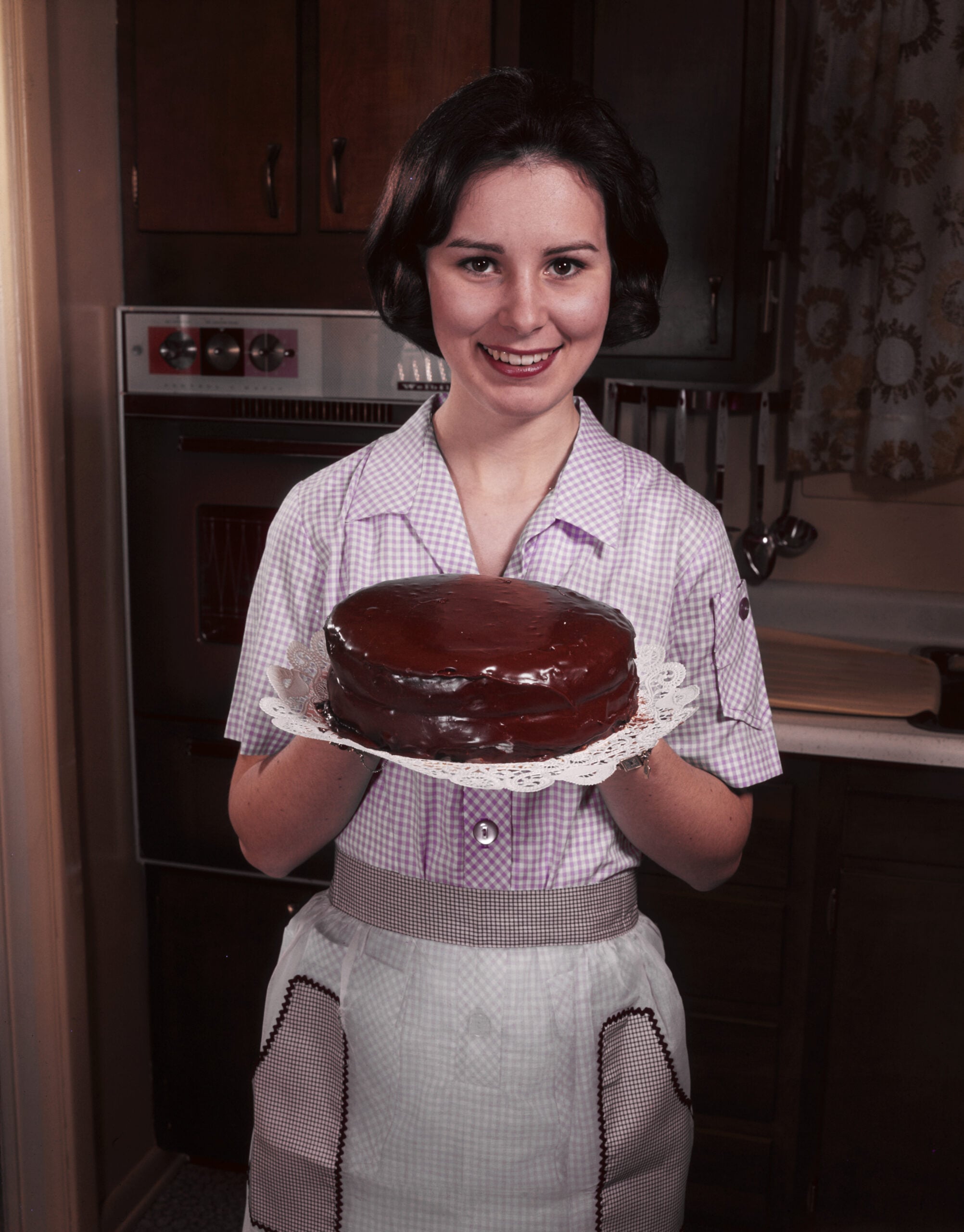 A woman in a checkered blouse and apron smiles while holding a chocolate cake on a doily in a vintage kitchen.