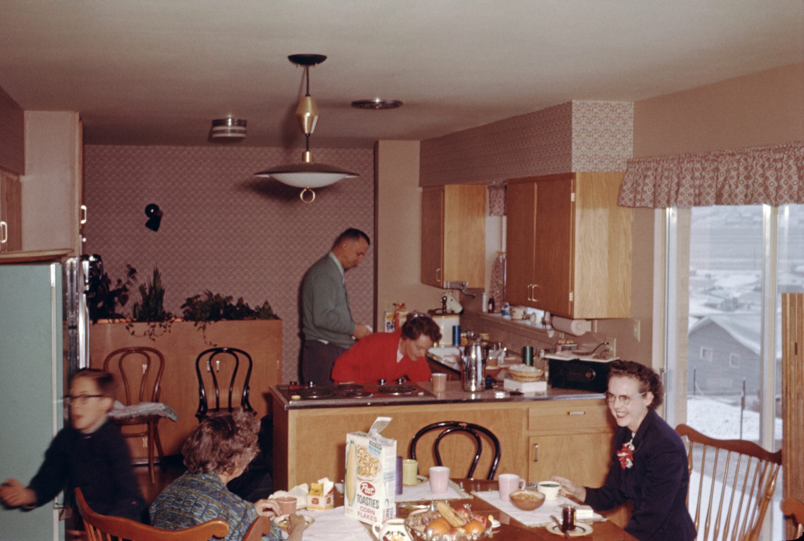 1950s family kitchens. Four people gather in a retro kitchen; two sit at a table with breakfast items, while two others prepare food at the counter. Sunlight comes through a large window, revealing a snowy neighborhood outside.