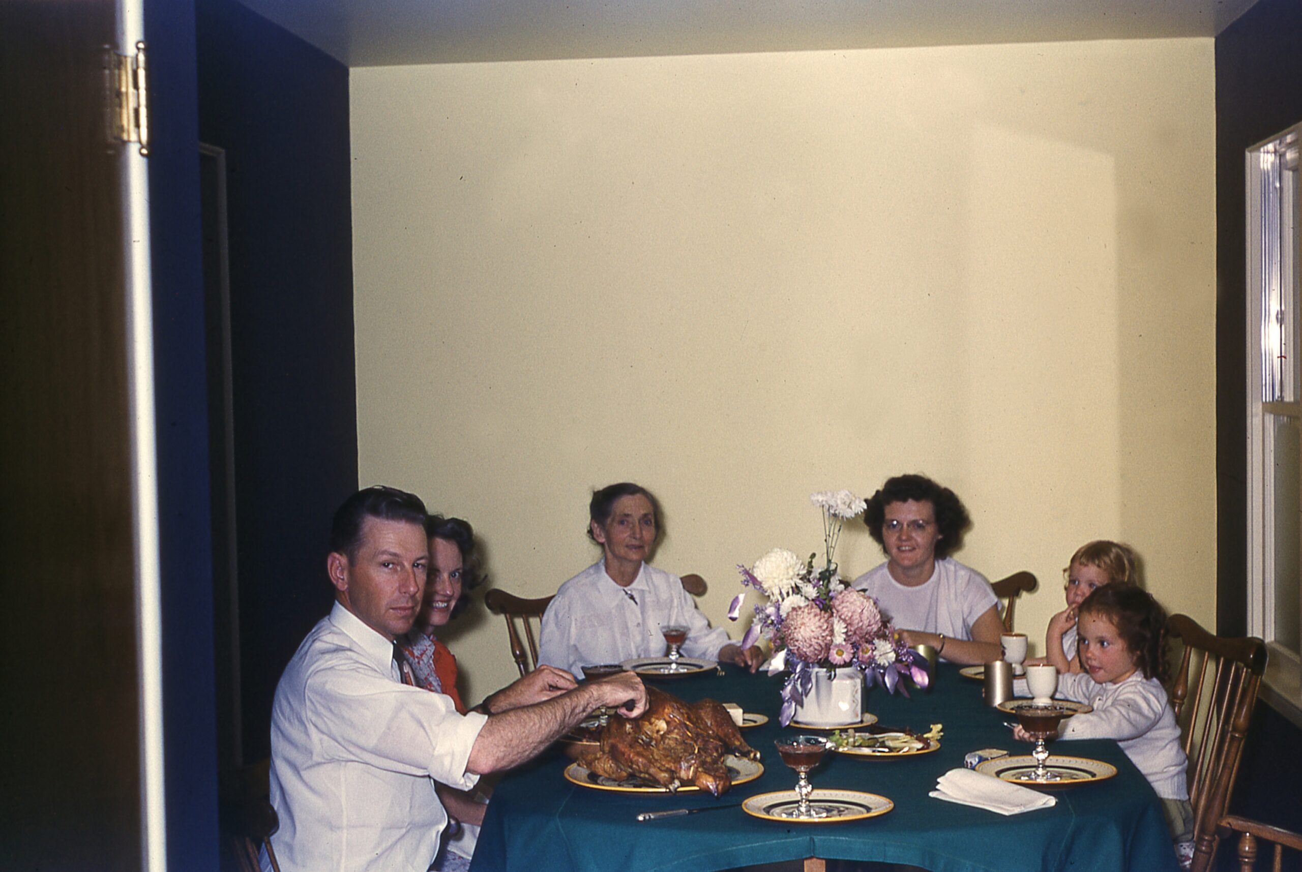 A family of six sits around a table with a roast turkey, plates, and flowers in the center, sharing a meal in a softly lit, cozy dining room.